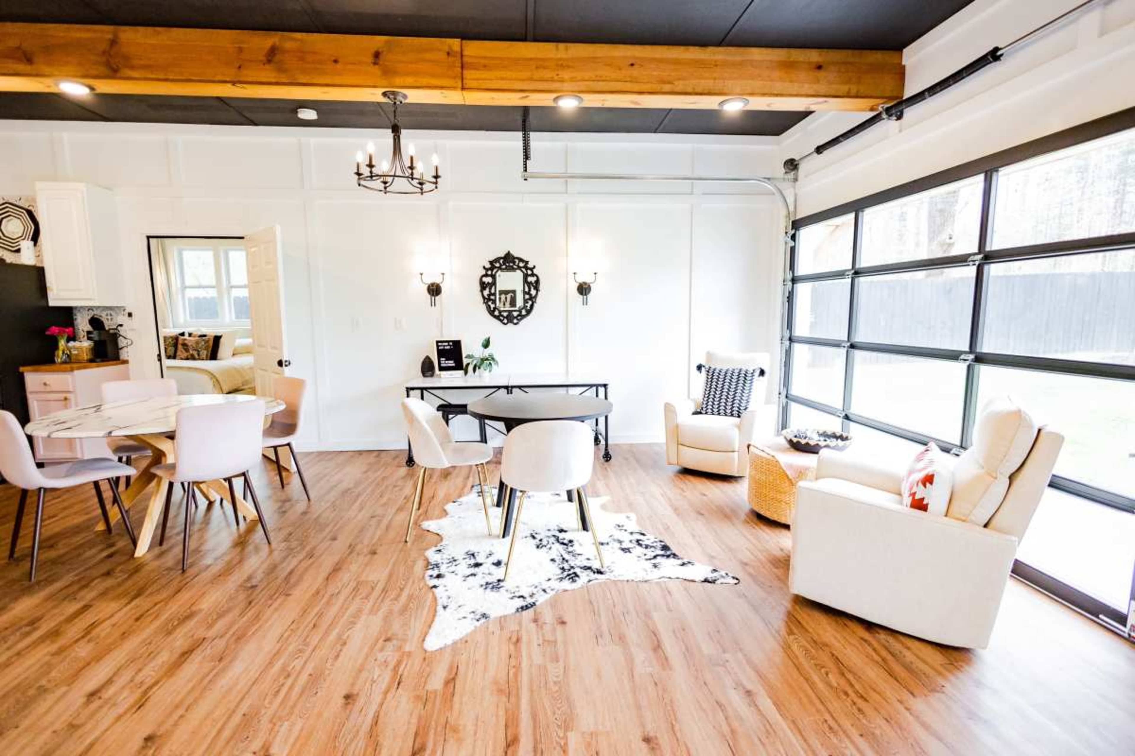 The image shows a modern interior space featuring a dining area with a round table, several chairs, a living area with two armchairs, and large glass doors that open to the outside.