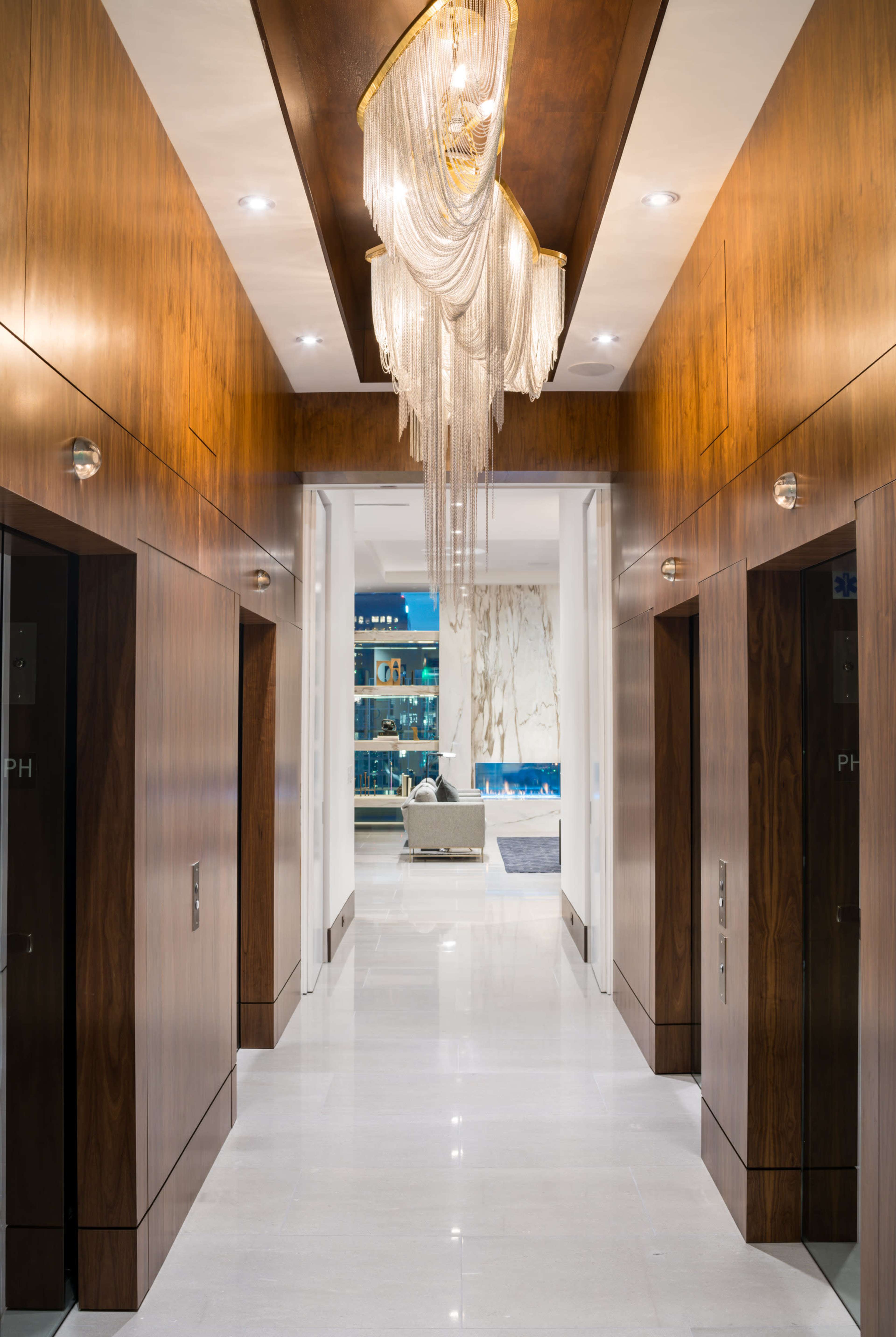 The image shows a luxurious hallway with wooden paneling, two elevators on the sides, and a decorative chandelier hanging from the ceiling.