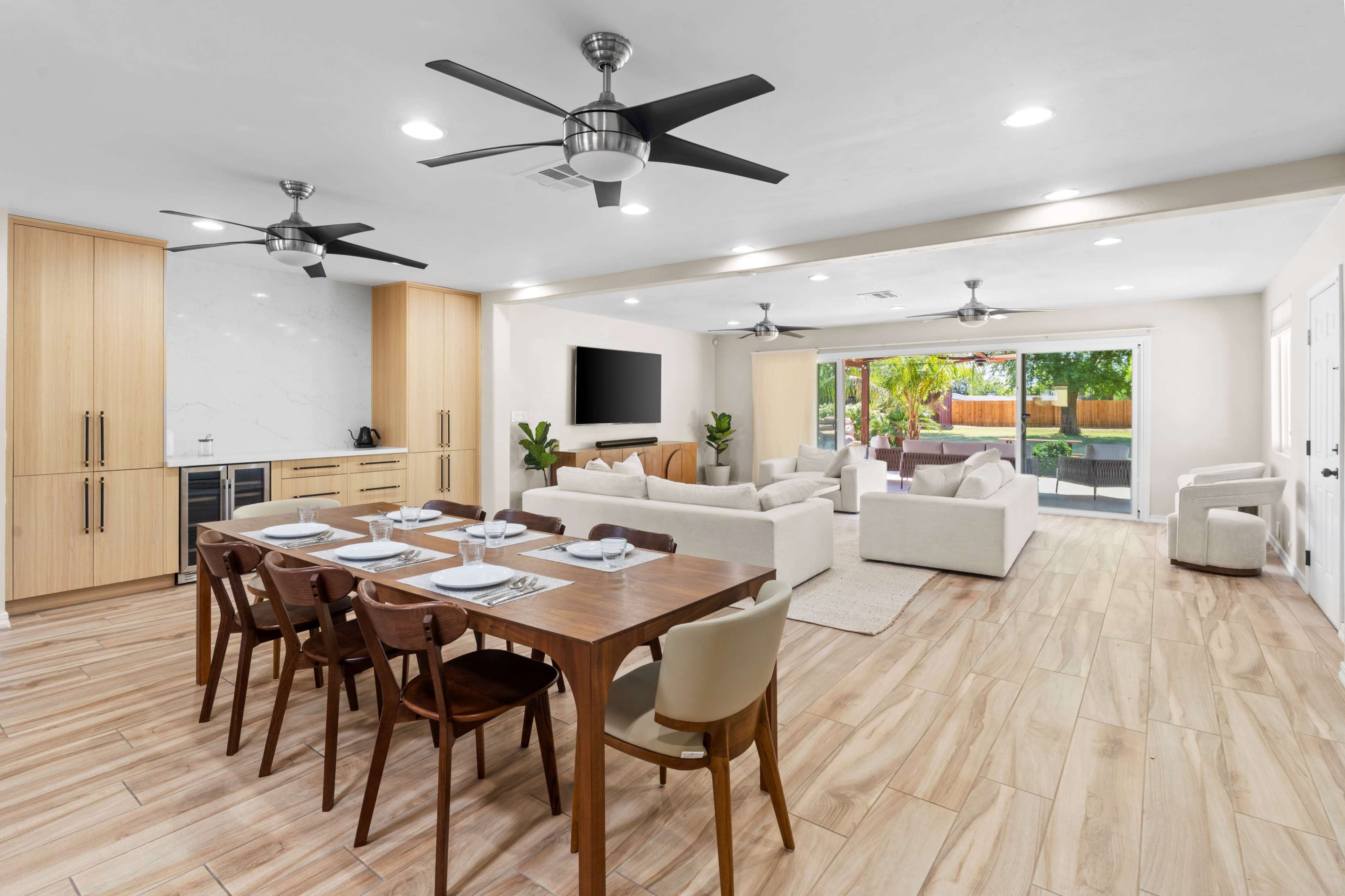 The image shows an open-concept living and dining area featuring a wooden dining table set for six, modern furniture, and large sliding glass doors leading to an outdoor space.