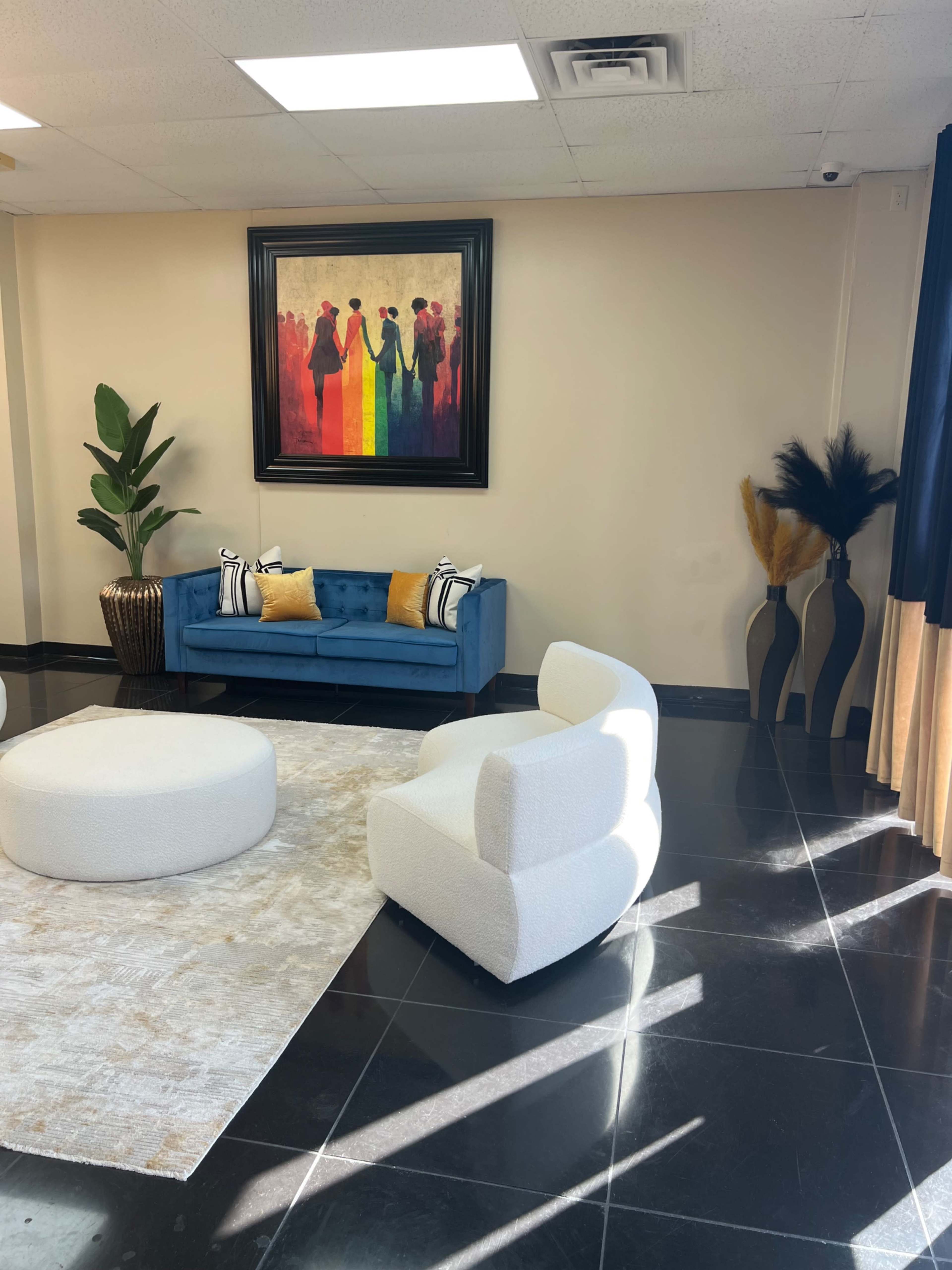 A modern interior space featuring a blue sofa with yellow and black pillows, a round white ottoman, and decorative plants near a large colorful painting on the wall.