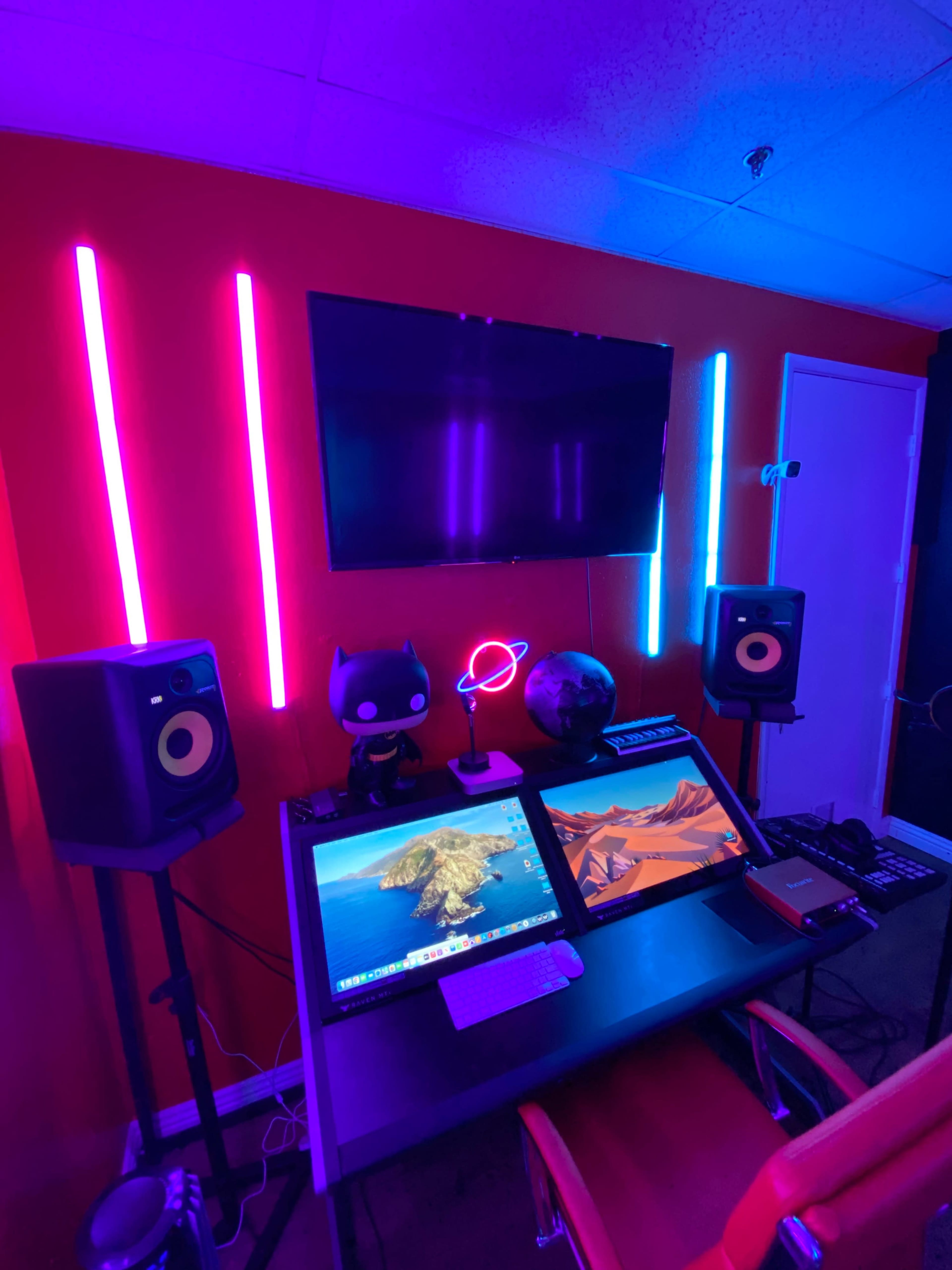 A modern workspace features dual monitors, colorful LED lights, a television on the wall, and decorative items like a globe and a figurine.