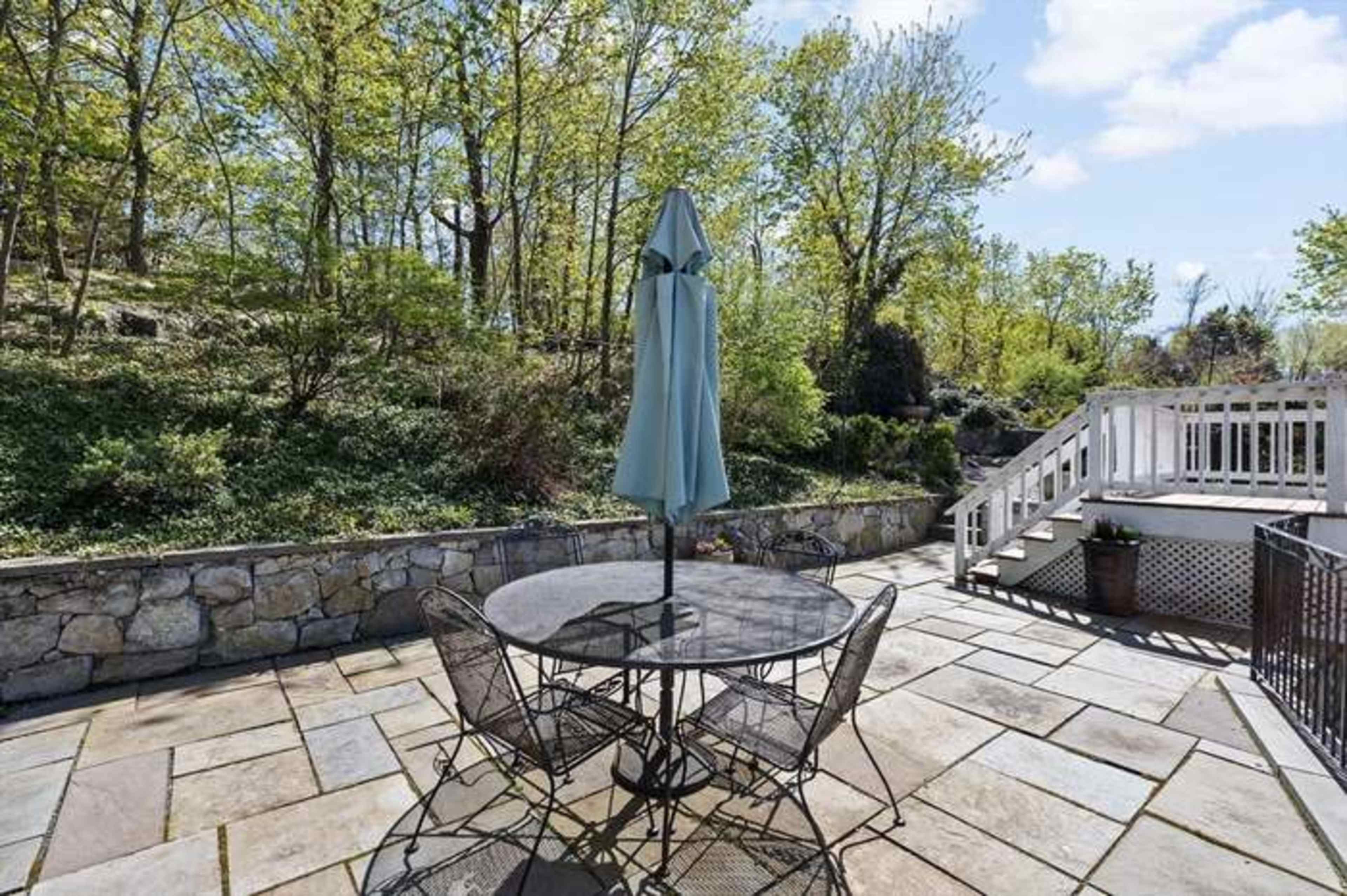 A stone patio features a circular table with four black metal chairs, accompanied by a light blue umbrella, and is surrounded by greenery and a staircase leading to a wooden deck.