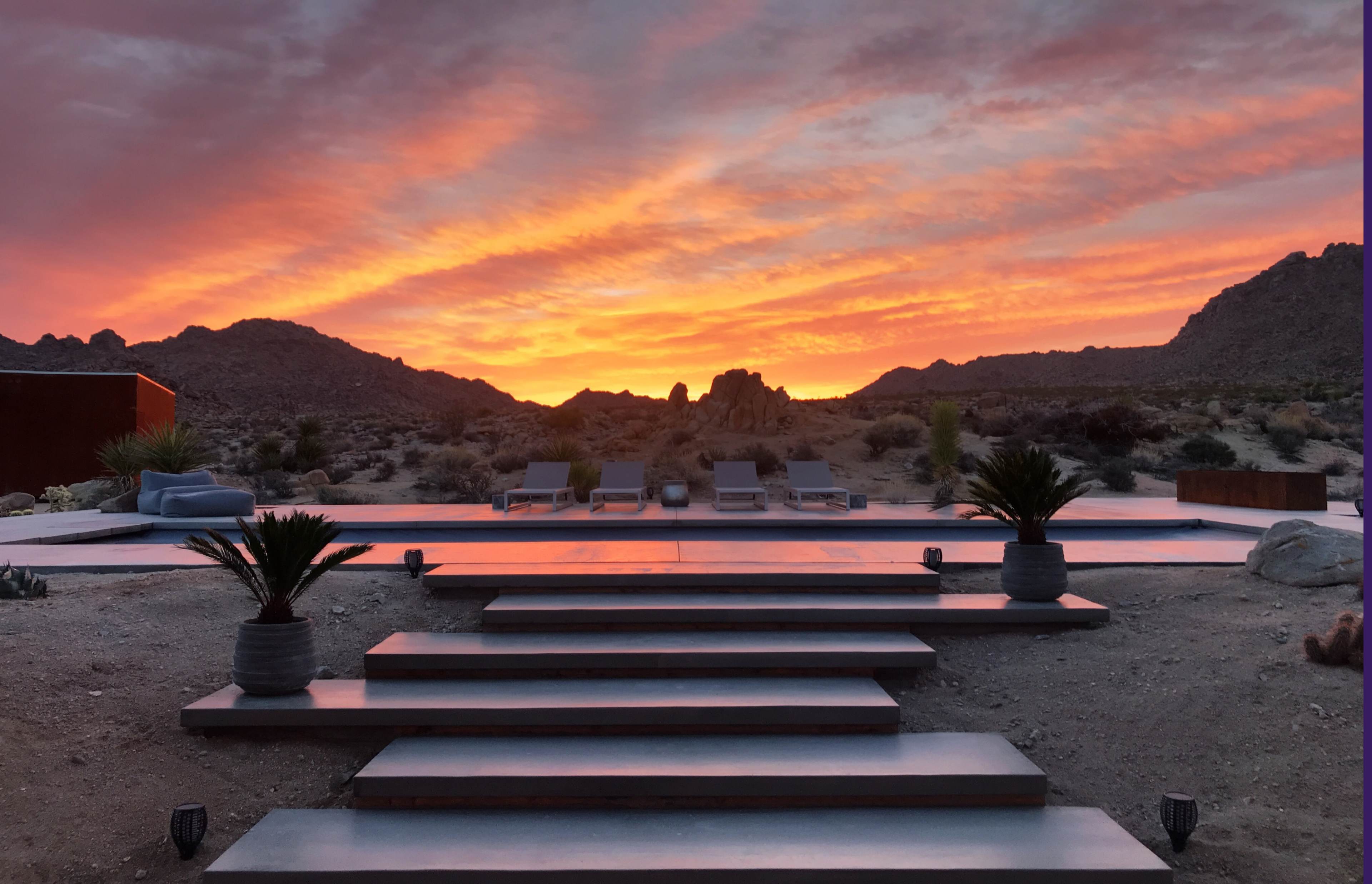 A desert landscape with stairs leading up to a modern patio, framed by rocky mountains and a vibrant sunset sky.