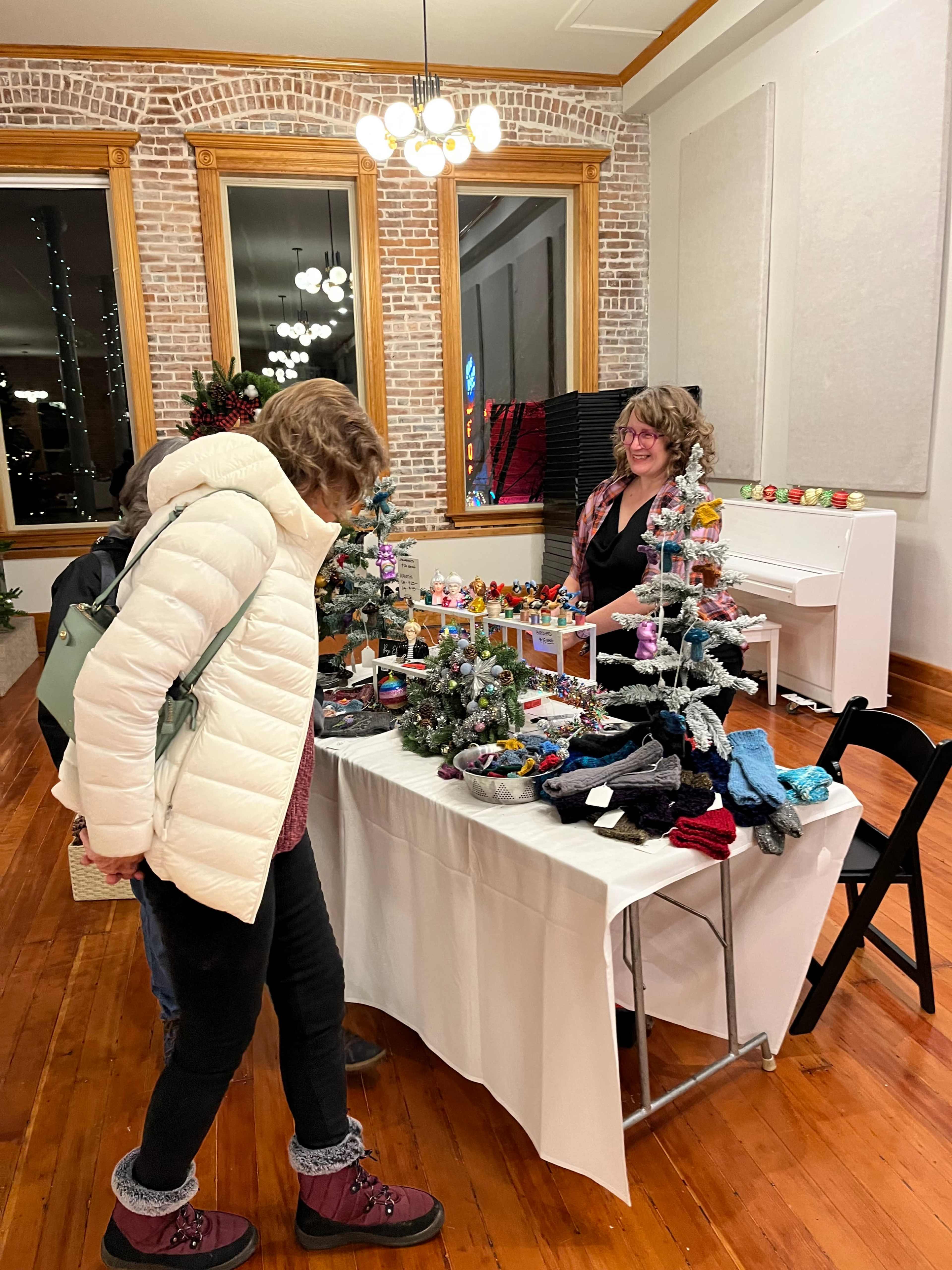 A vendor stands behind a table displaying handmade knitted items while two customers examine the products in a warmly lit indoor market setting.