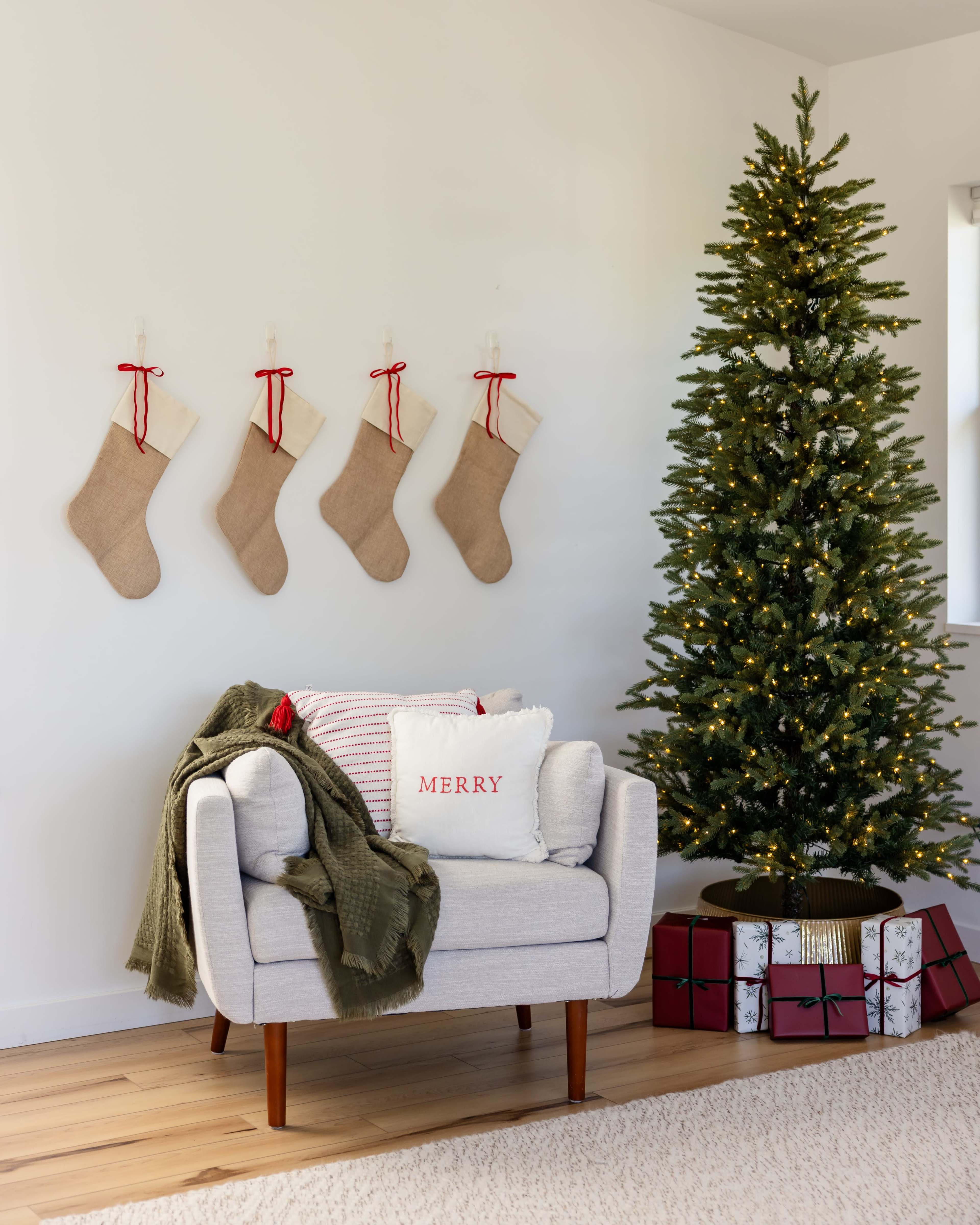A modern living room features a decorated Christmas tree, festive stockings hanging on the wall, and a cozy armchair with decorative pillows.