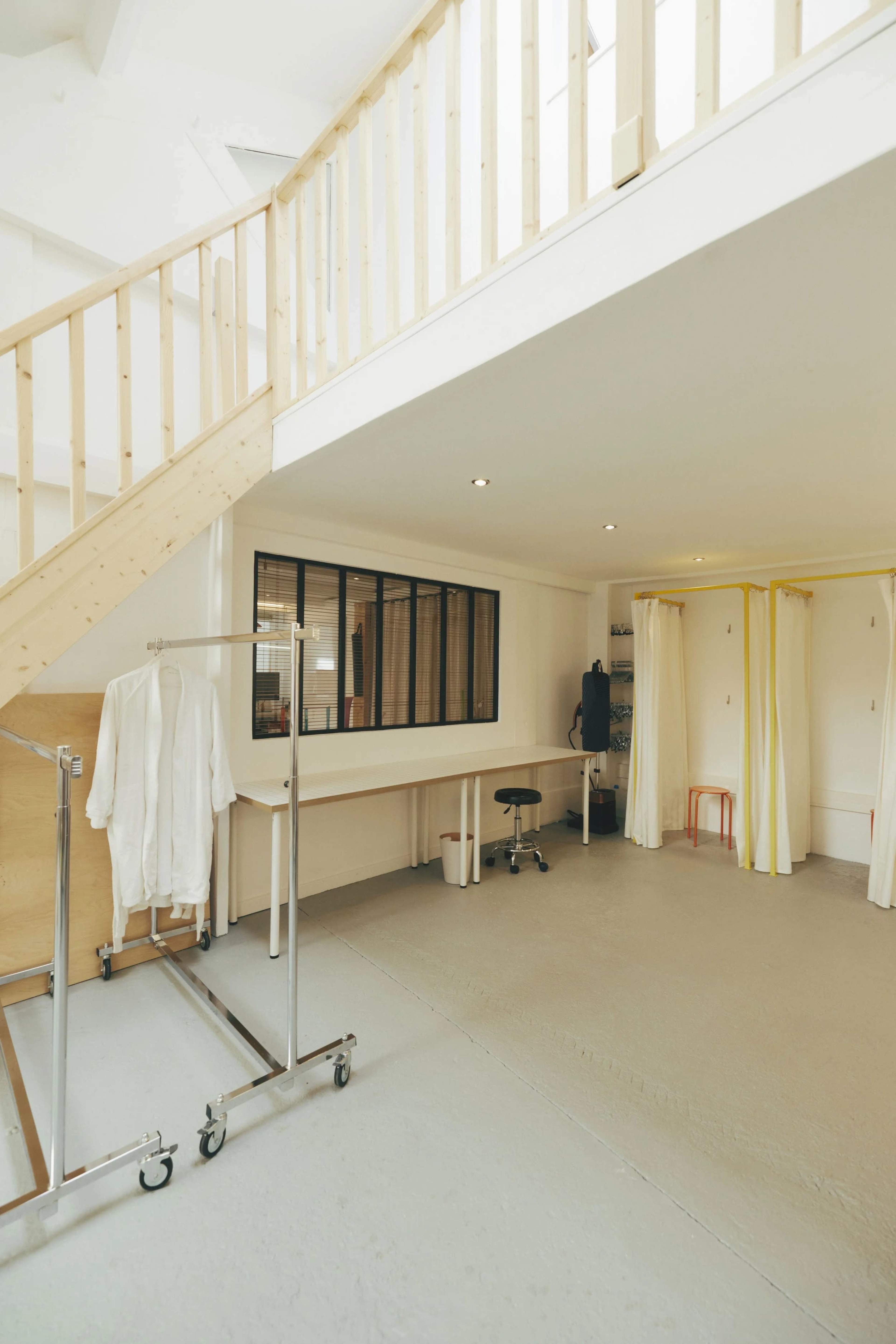 The image shows a spacious interior with a staircase, a large window, a table, a rolling clothing rack, and two curtain partitions.