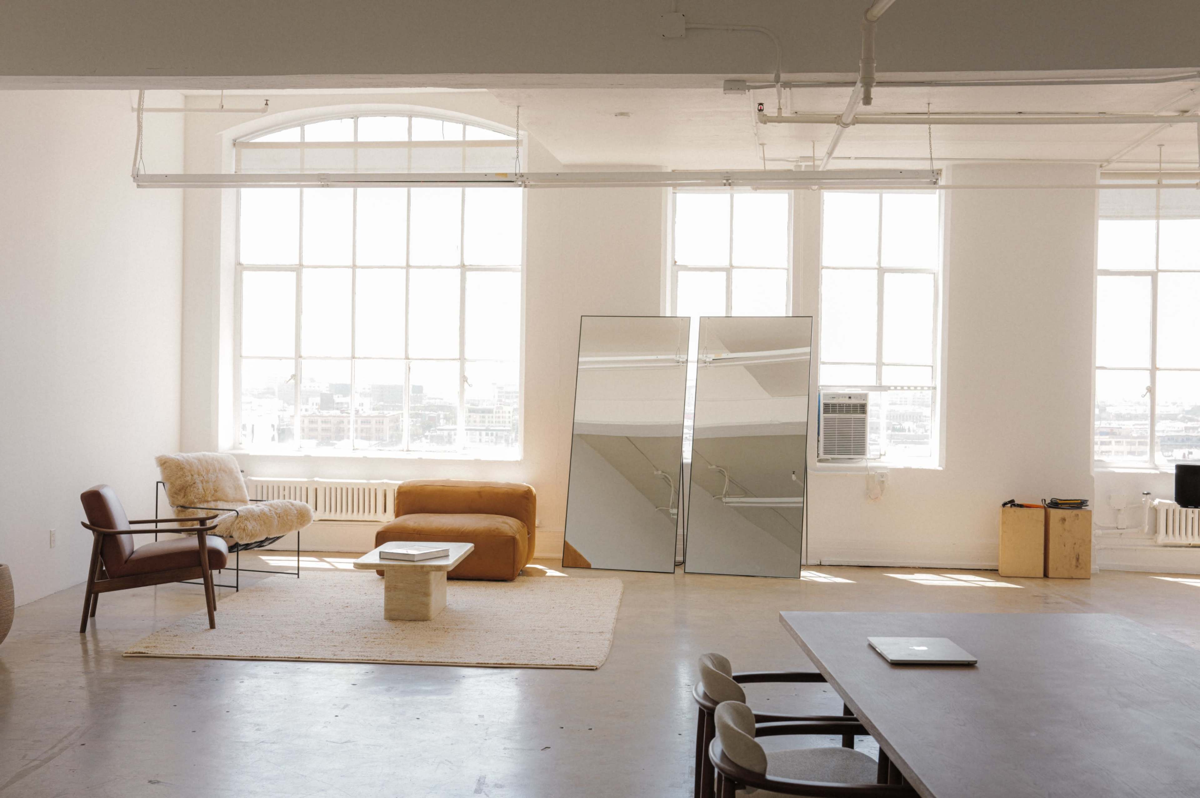 The image depicts a spacious, bright room with large windows, featuring a mix of modern furniture including a sofa, a chair, a table, and two large mirrors.