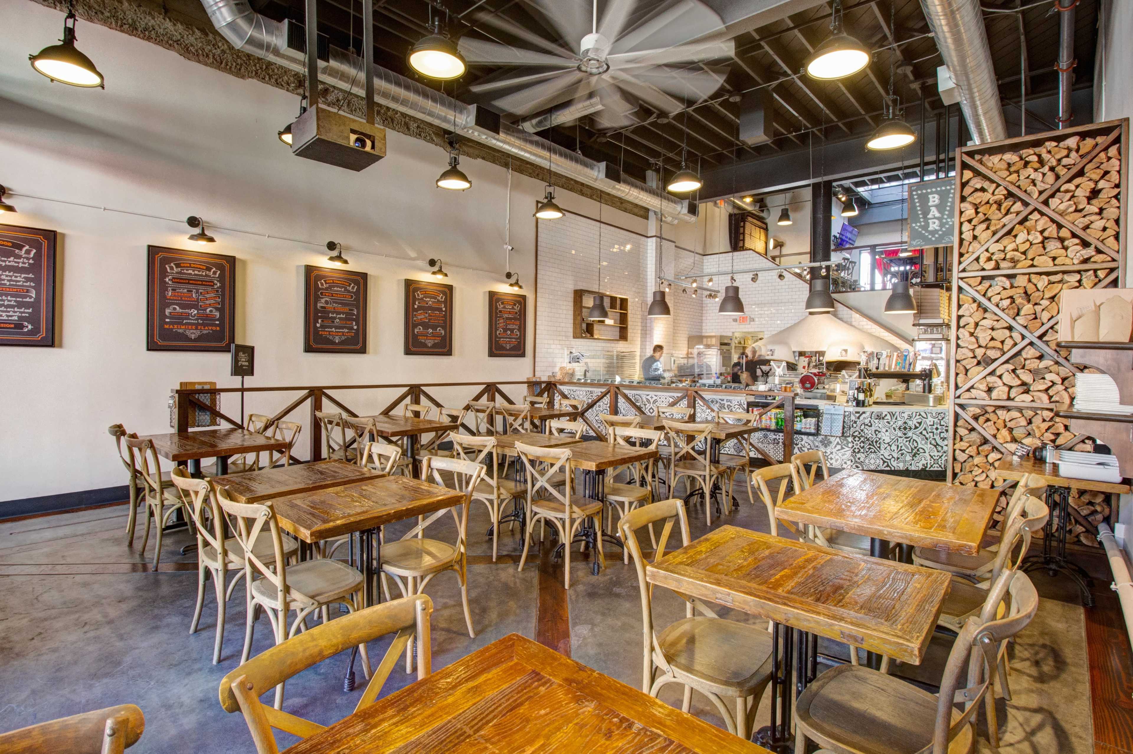 A spacious restaurant interior featuring wooden tables and chairs, with a visible kitchen area in the background and menus displayed on the walls.