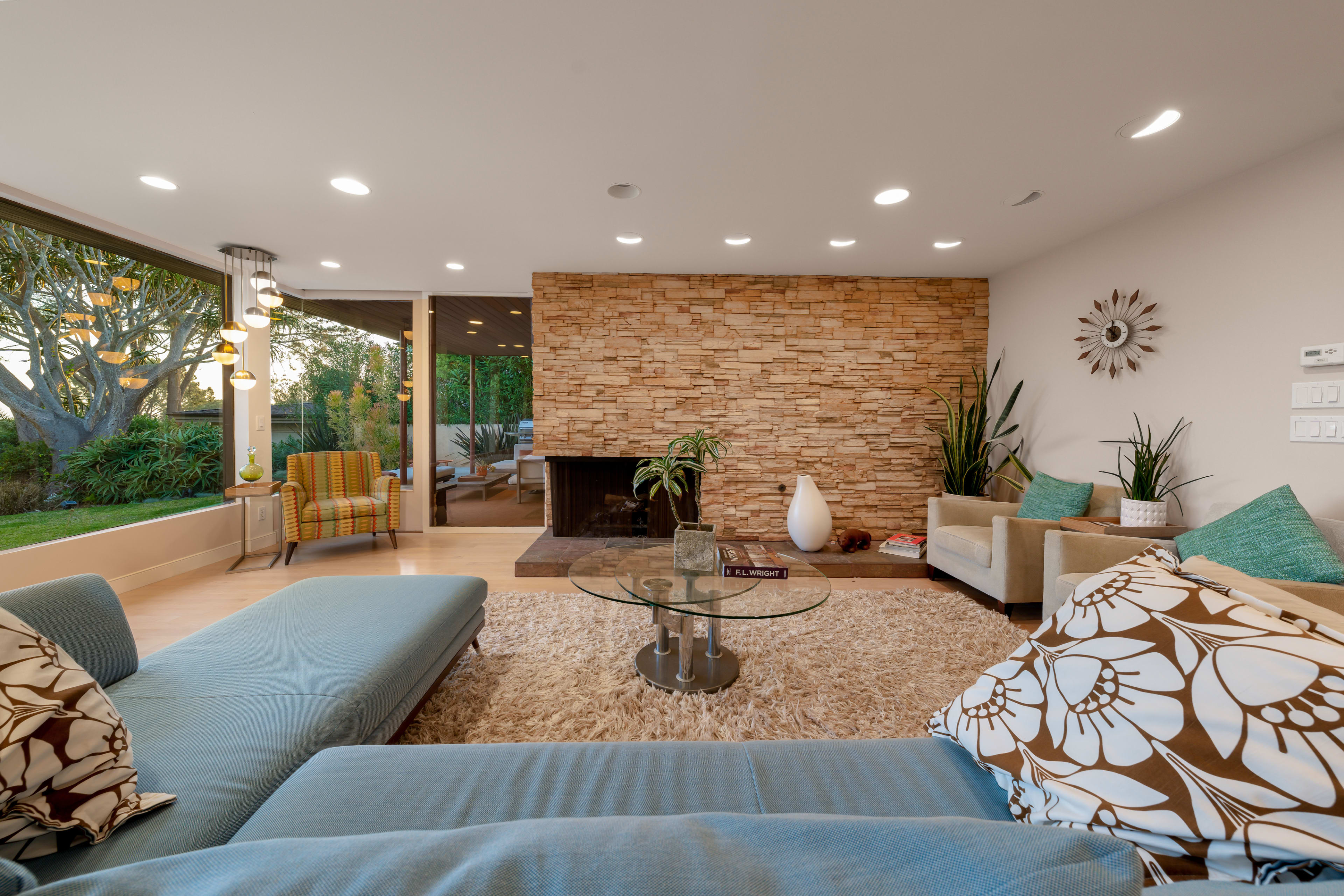 A modern living room features a stone fireplace, a glass coffee table, and seating arrangements with decorative pillows, set against large windows overlooking a garden.
