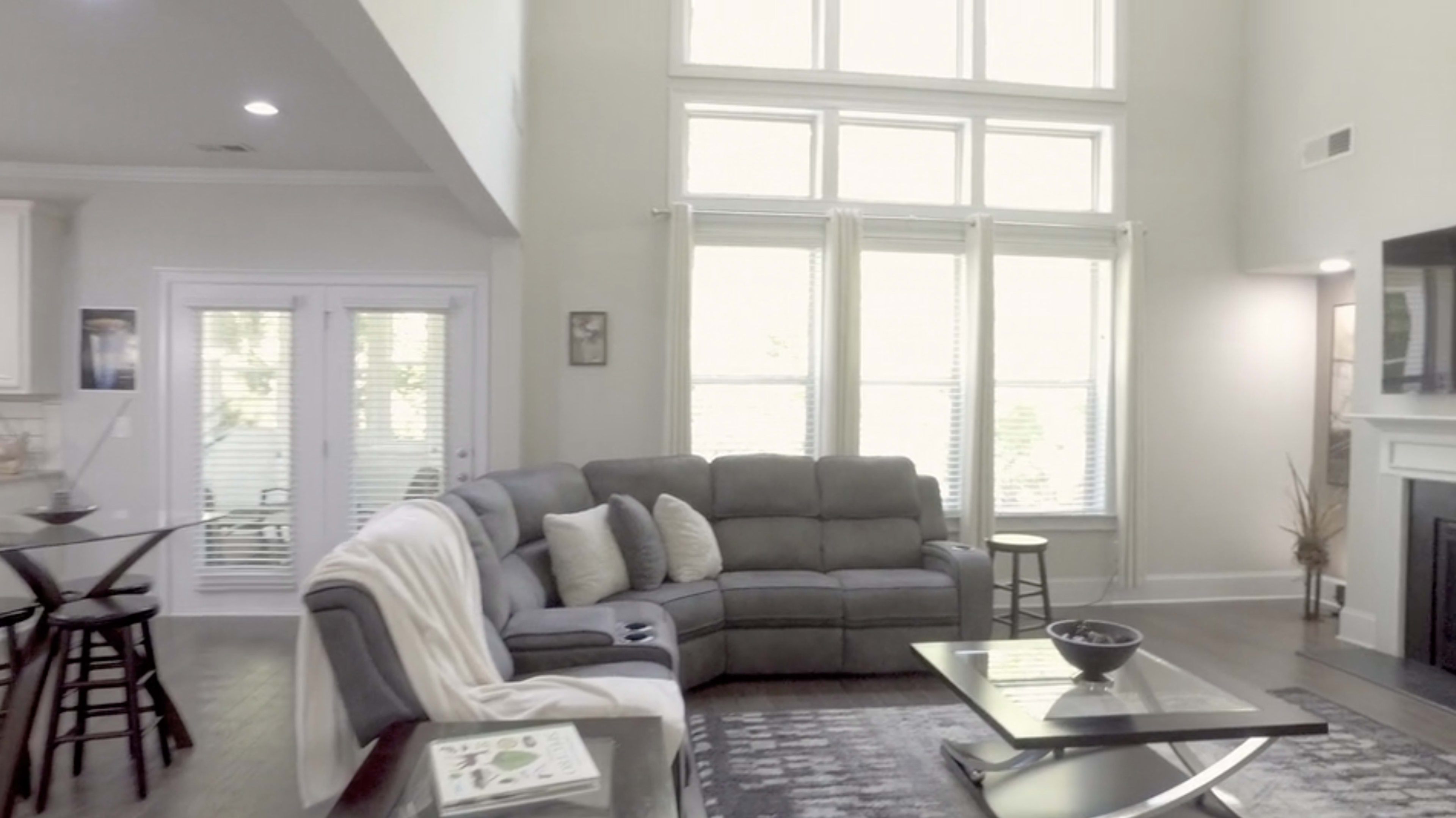 A spacious living room with a gray sectional sofa, large windows allowing natural light, and a dining area visible in the background.