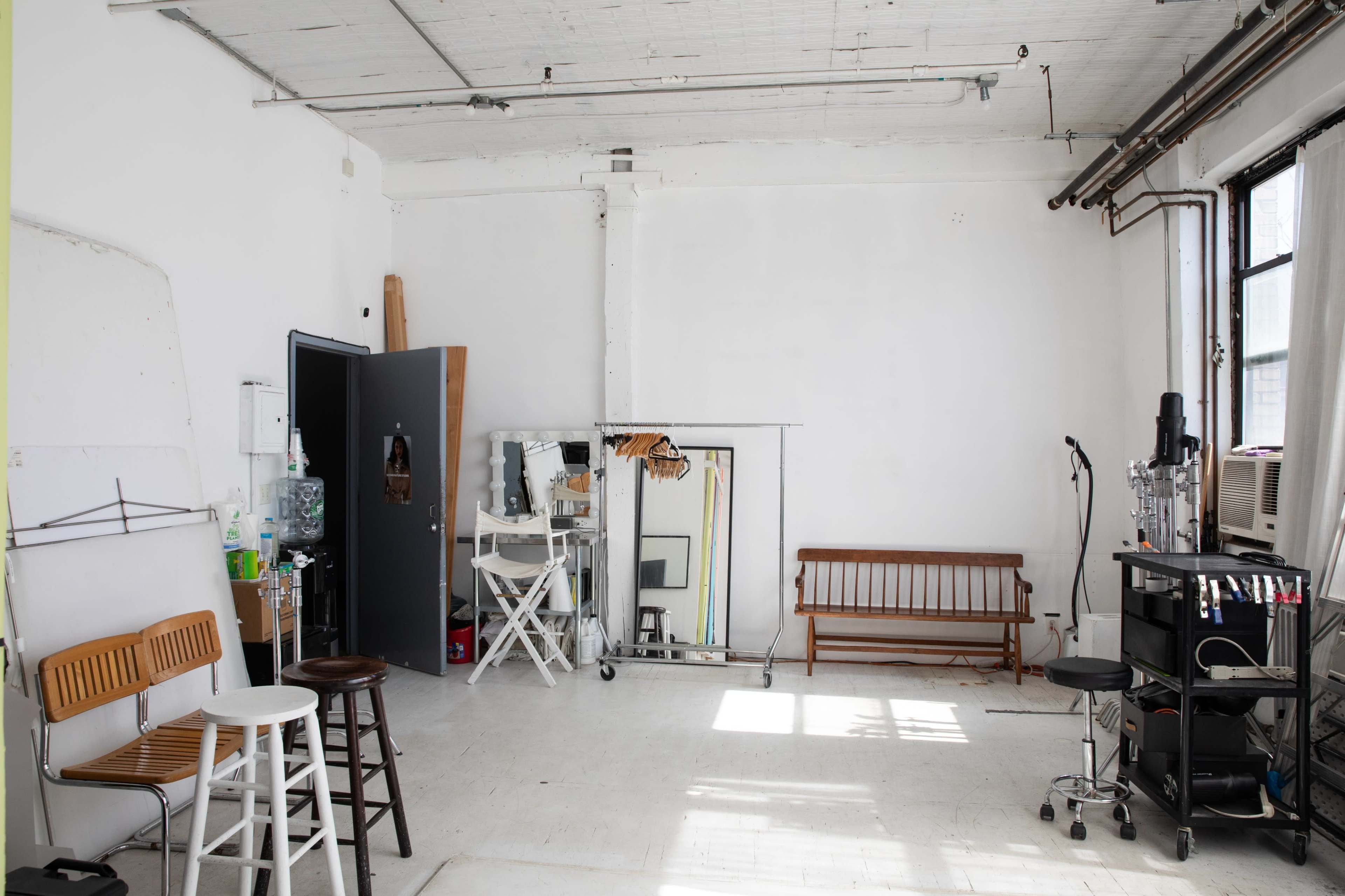 The image shows a sparse, well-lit room with white walls, featuring a few chairs, a mirror, a small cart, and a window air conditioning unit.