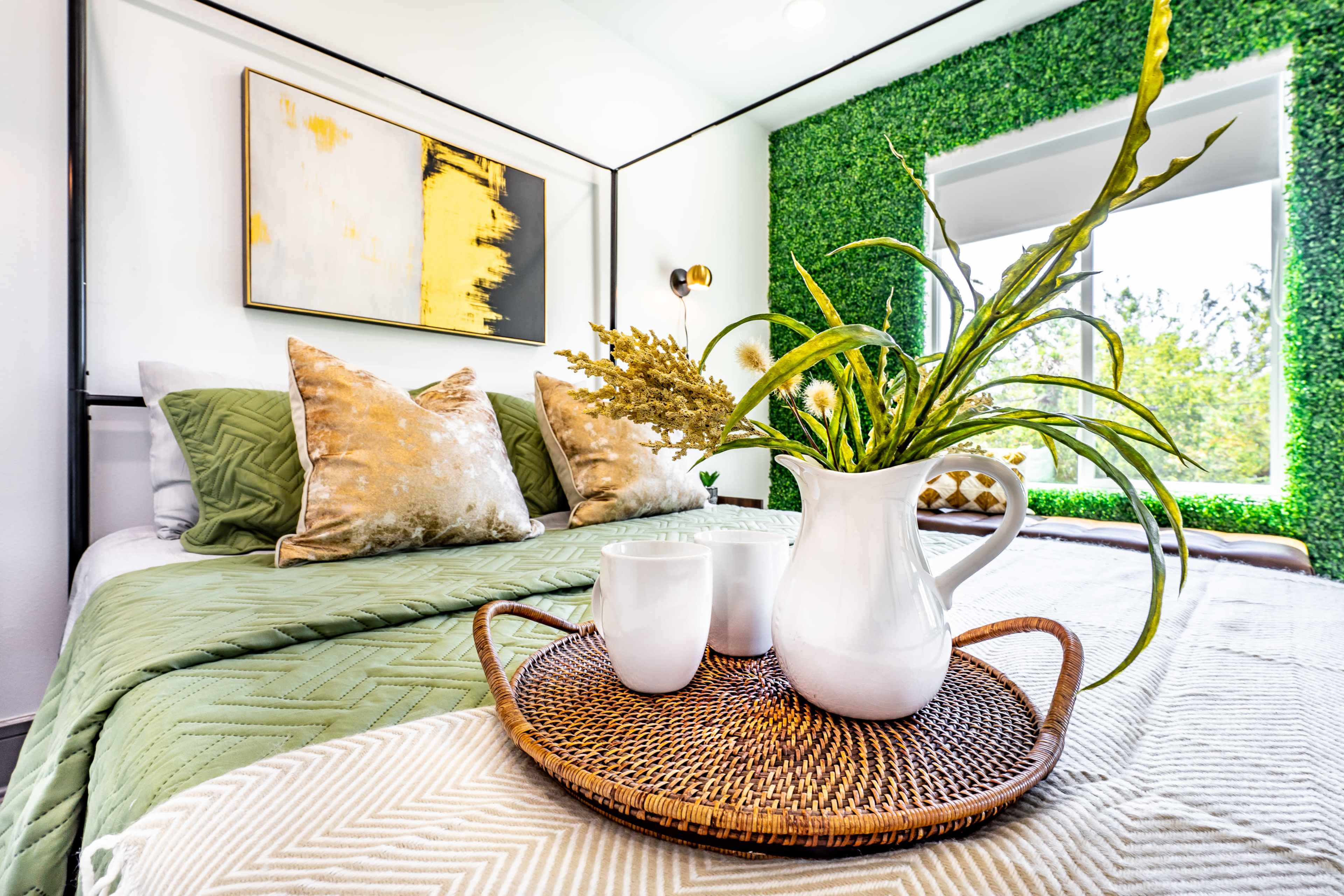 A neatly arranged bedroom with green decor, featuring a bed with decorative pillows and a tray of a pitcher and cups on a table.