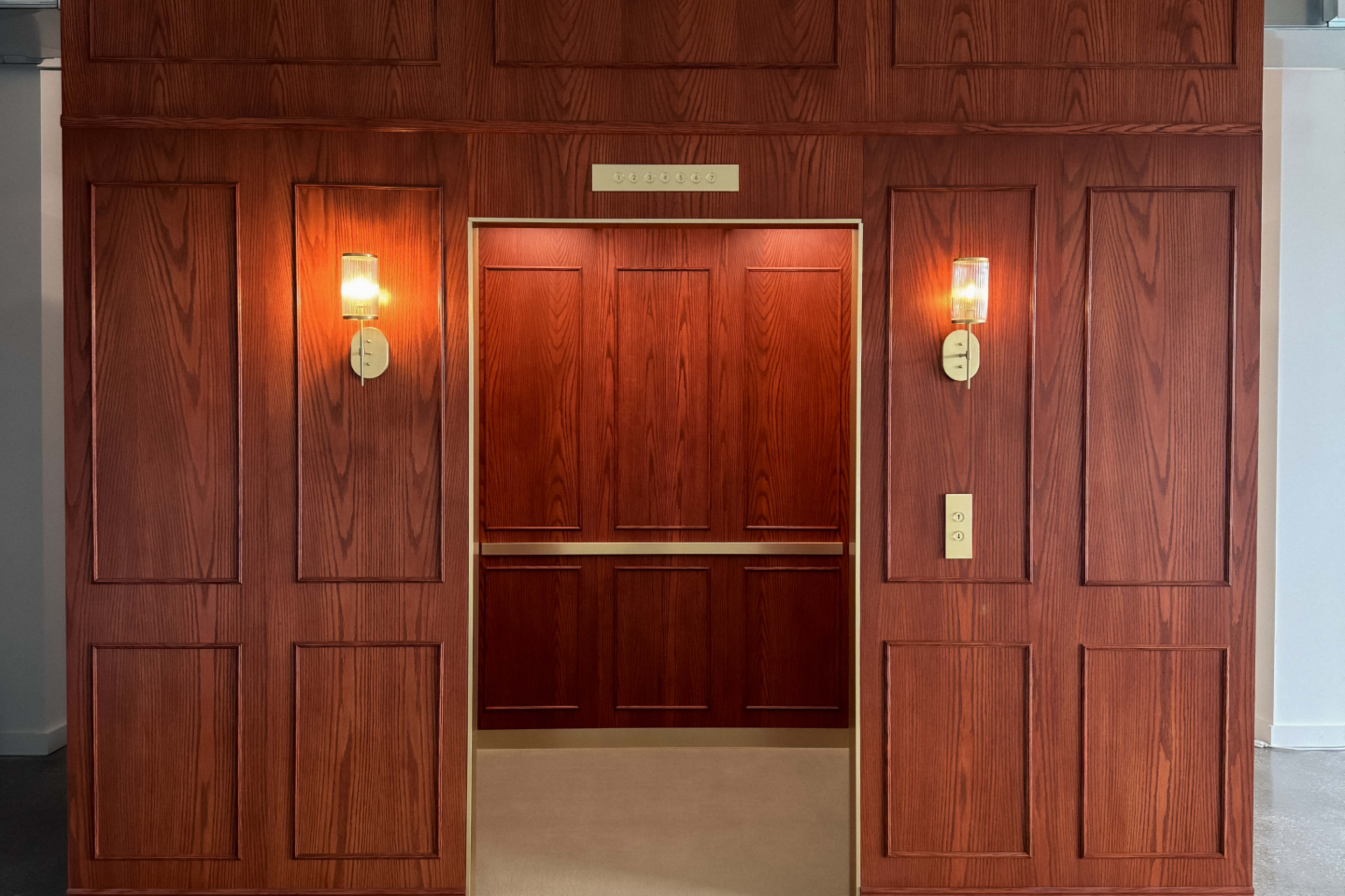 The image shows a wooden-paneled elevator entrance with two wall-mounted lights flanking the open doors.