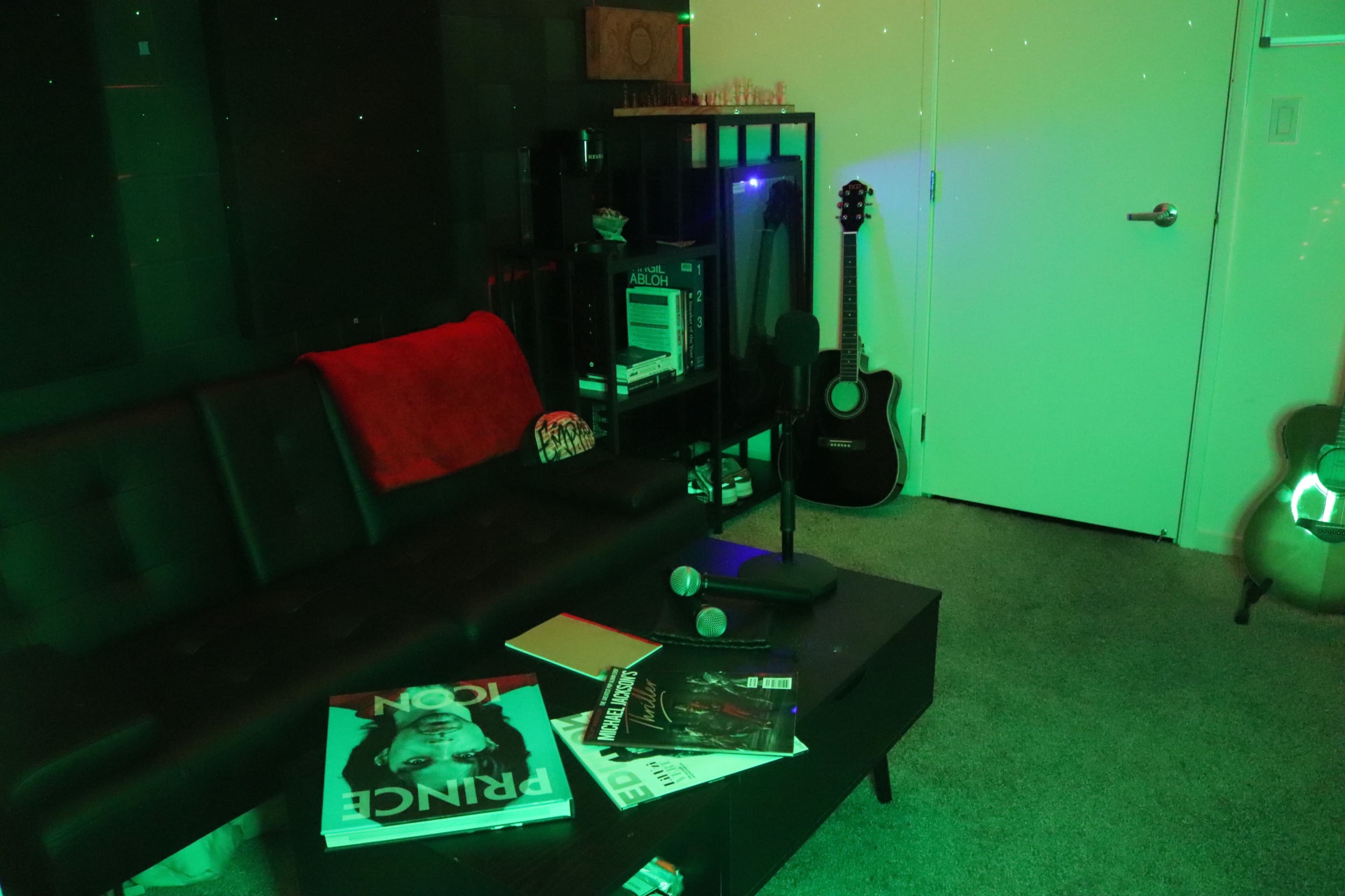 The image shows a small room with a black sofa, a coffee table covered with magazines, and musical instruments, including an acoustic guitar, against a wall.