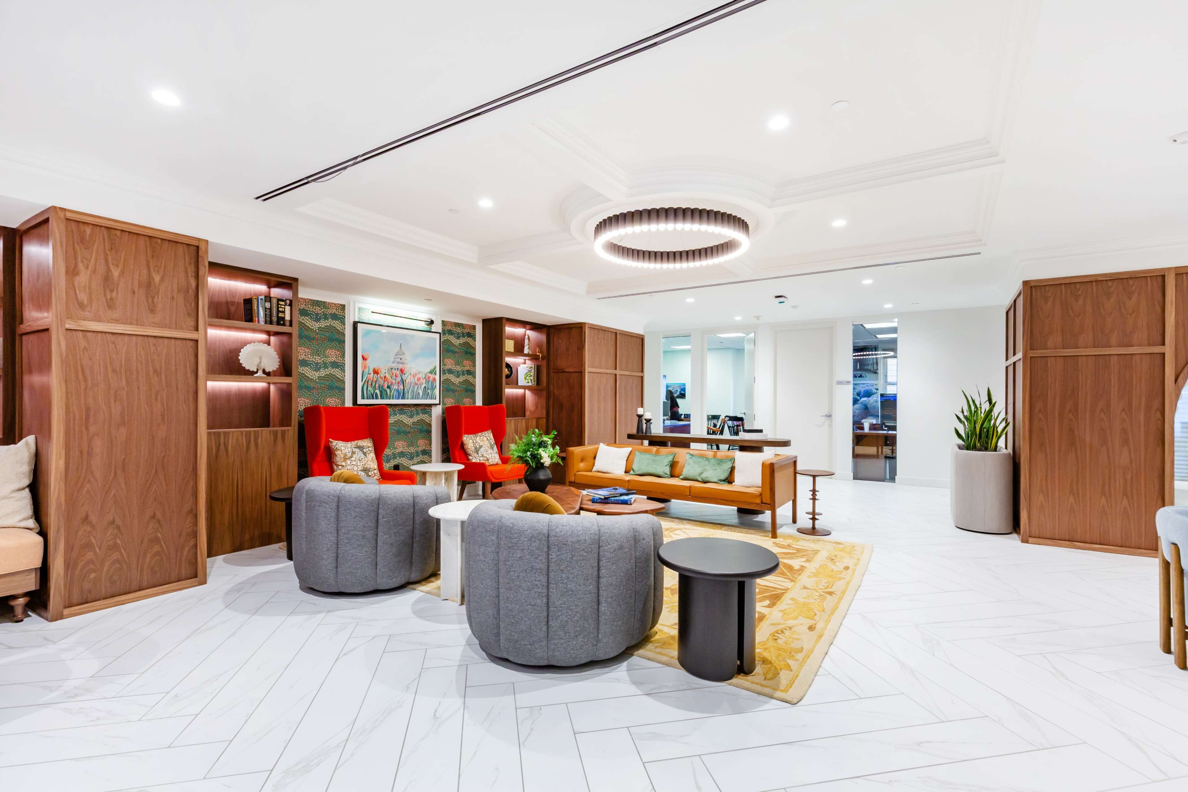 The image shows a modern lobby area featuring a seating arrangement with two red chairs, gray circular couches, a wooden coffee table, and decorative plants, all set against a white tiled floor.