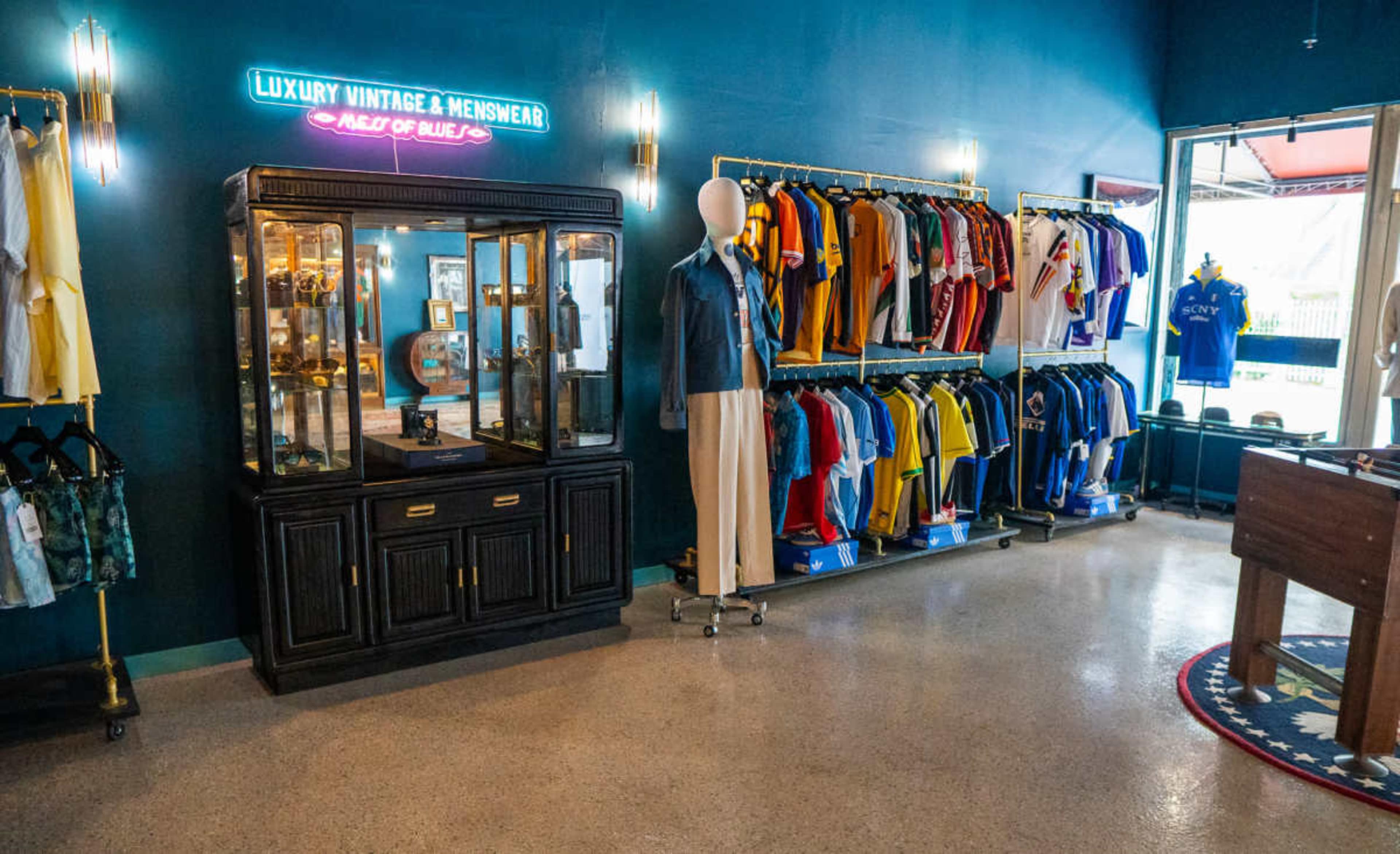 The image shows a retail space displaying vintage and menswear items, featuring a glass display cabinet and clothing racks filled with various colorful shirts and dresses against a blue wall.