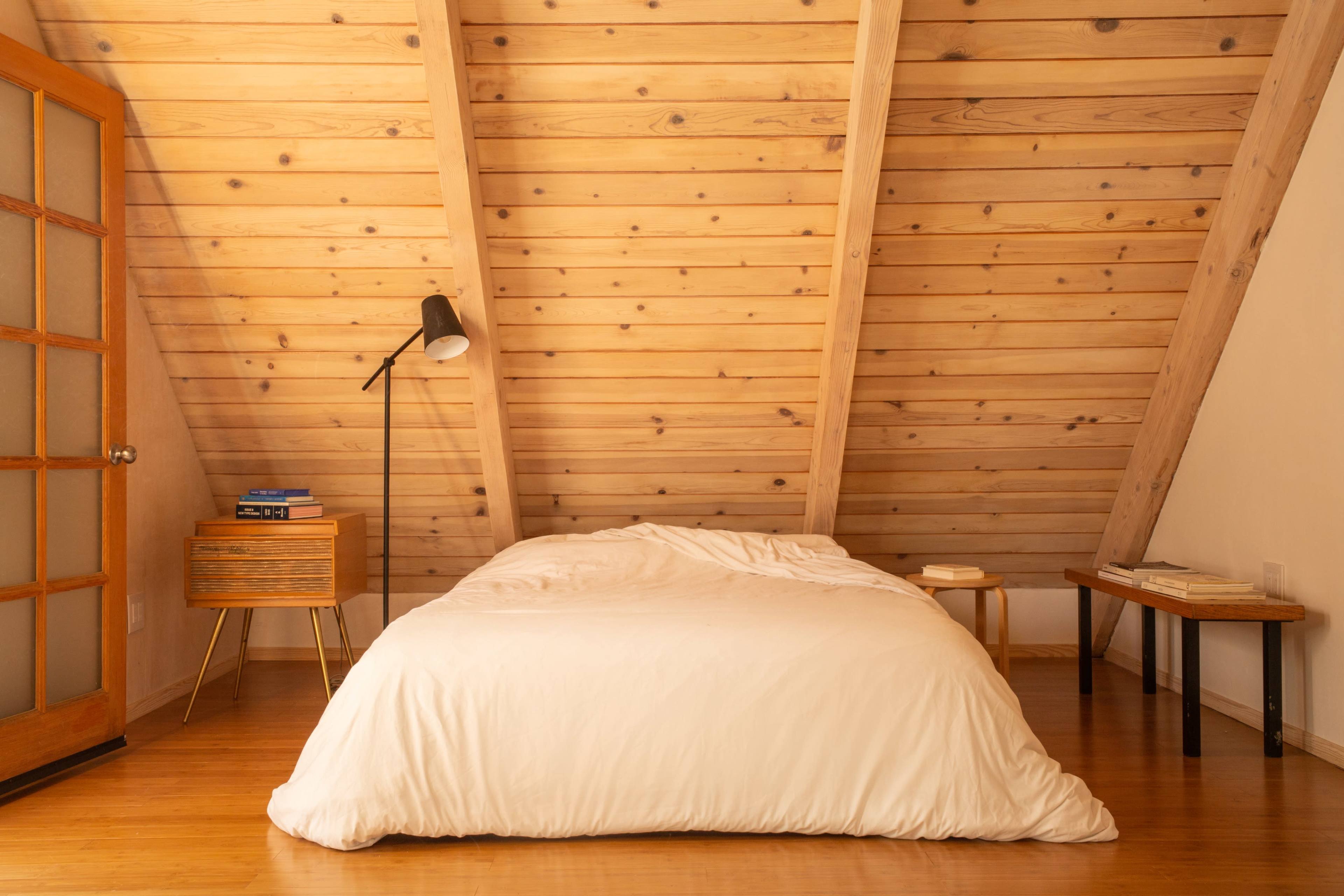 A simple bedroom features a bed with a white coverlet under a sloped wooden ceiling, accompanied by a bedside table and a floor lamp.