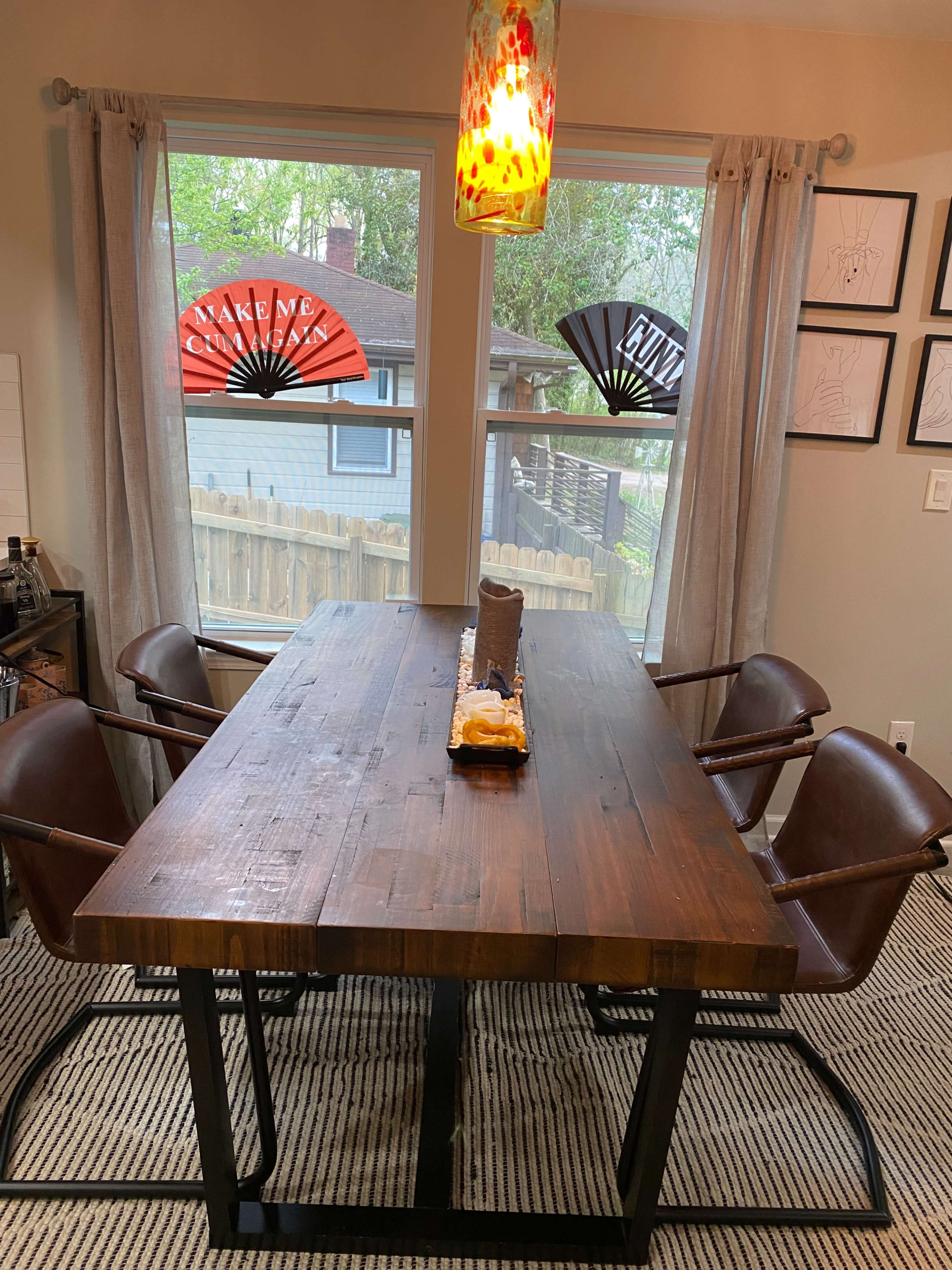 A wooden dining table with four leather chairs is set near two windows featuring decorative fans.