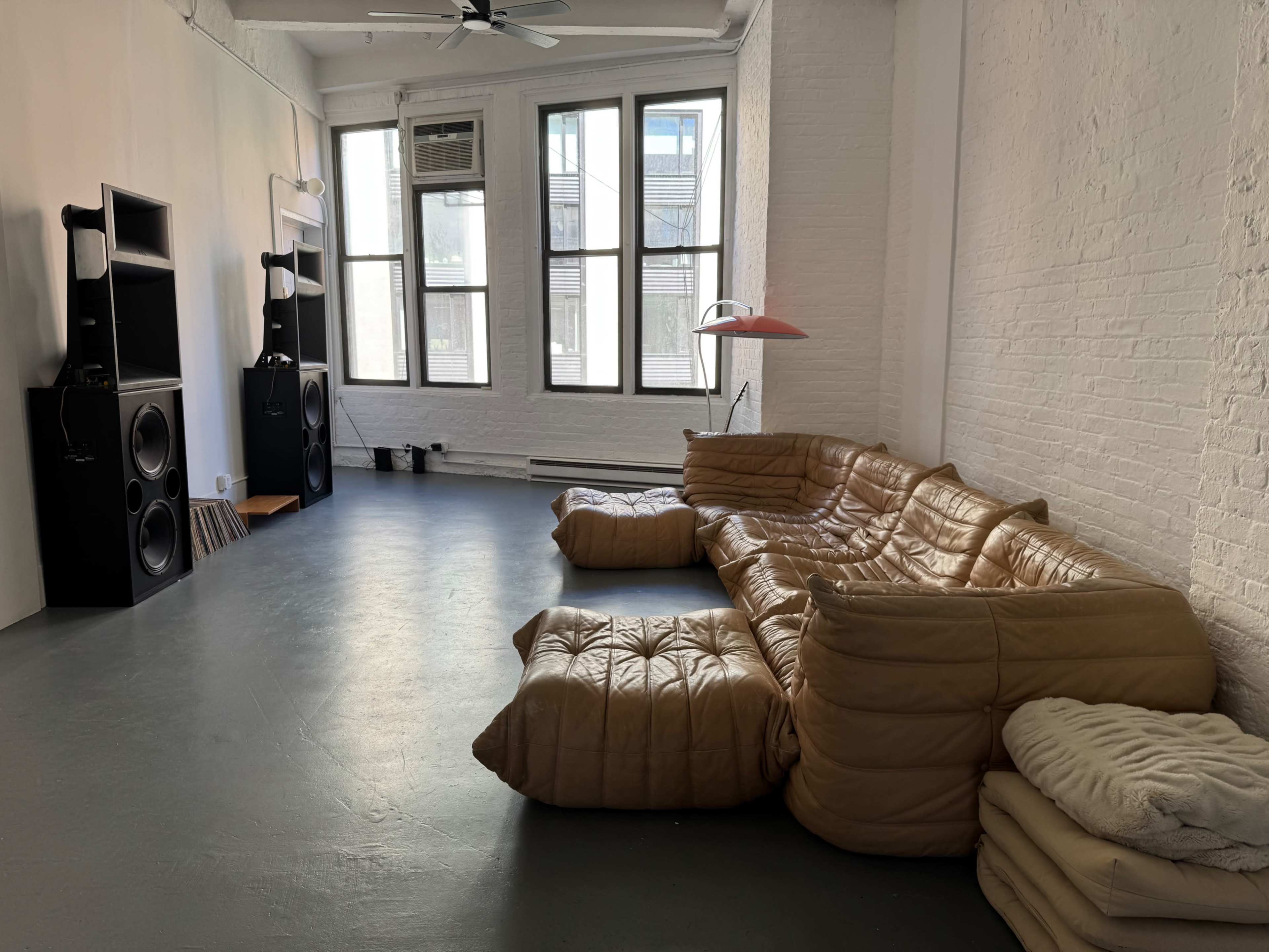 The image shows a spacious room with large windows, featuring a curved tan leather sofa, two large speakers, and a modern lamp.
