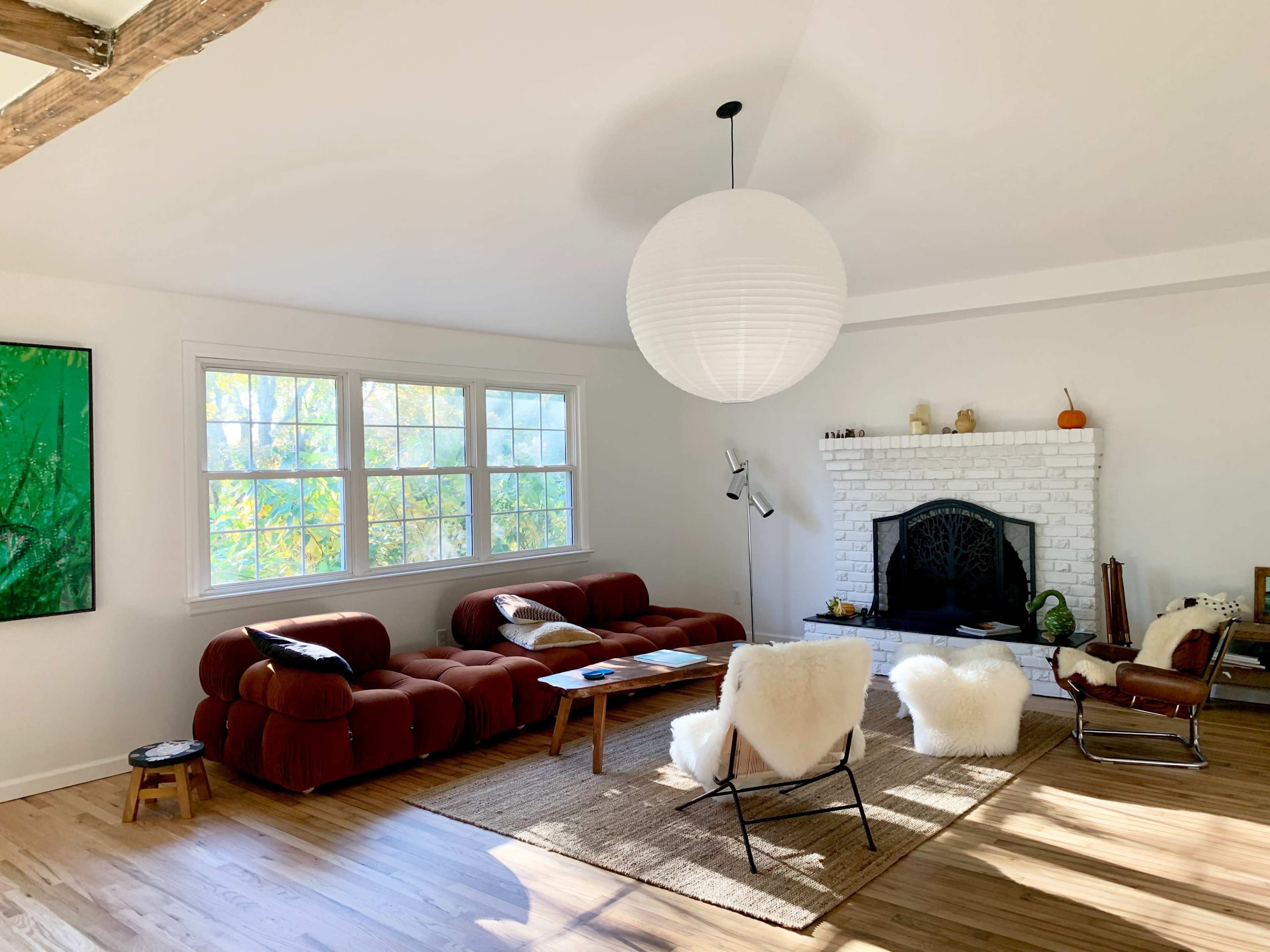 A modern living room features a brown sectional couch, white fireplace with a mantel, and a large pendant light, with sunlight streaming through the windows.