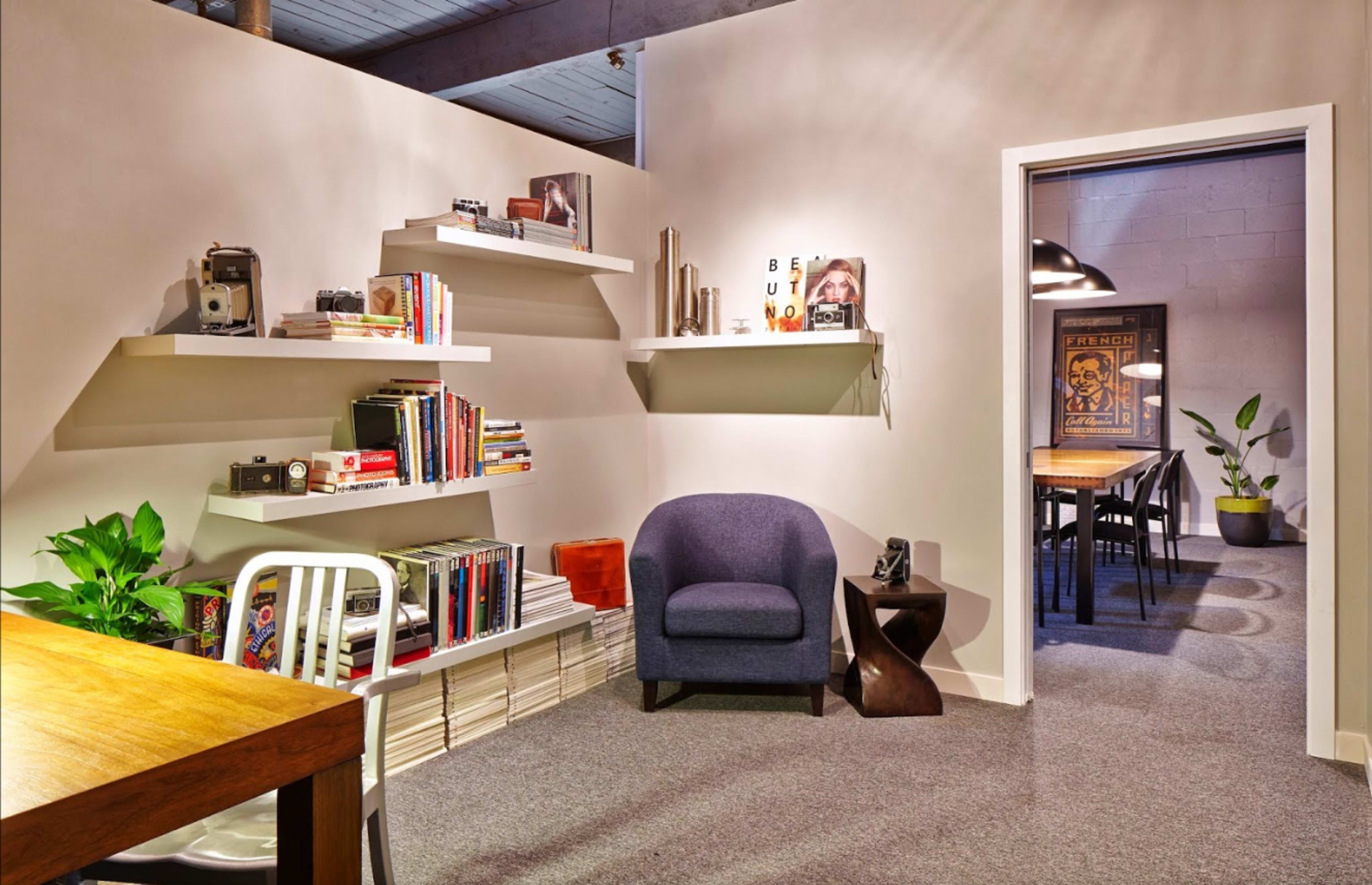 A corner of a well-lit room features a chair beside a wall with bookshelves filled with books and cameras, leading to a dining area visible through an open door.