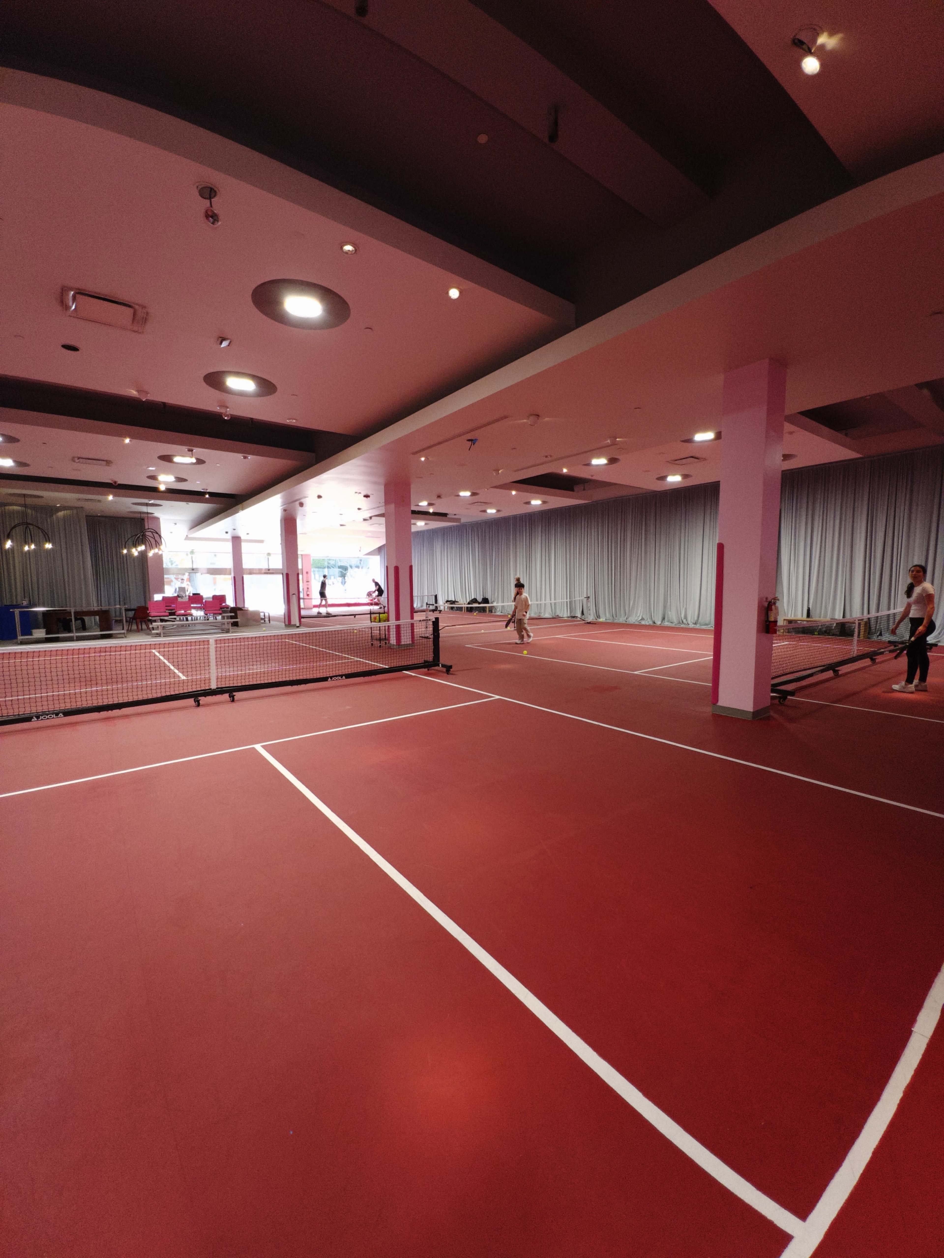 The image shows a spacious indoor tennis facility with a red court surface, featuring multiple courts separated by nets.