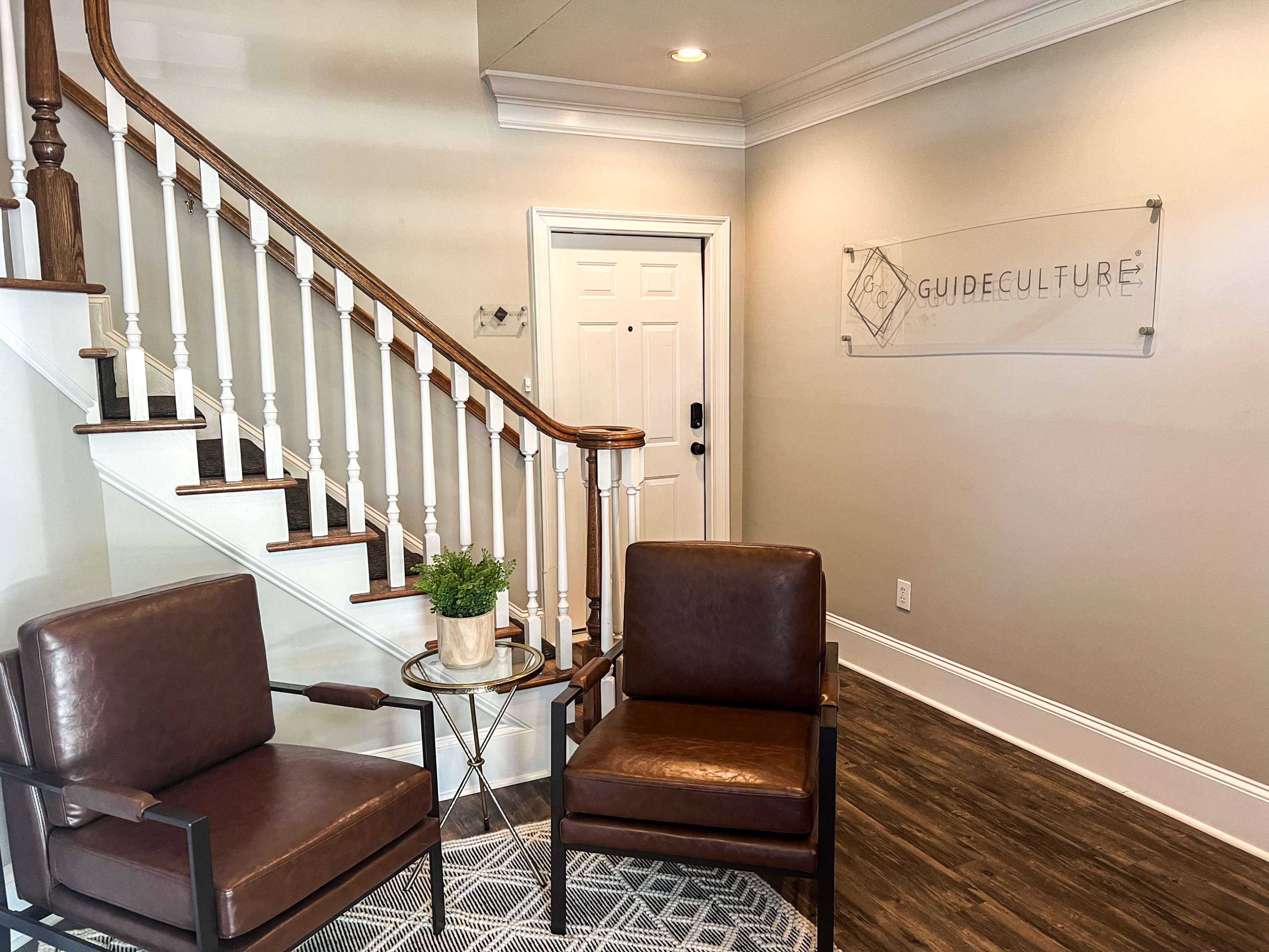 A waiting area with two brown chairs, a small round table, a potted plant, and a staircase leading to a door marked with a sign that reads "GUIDE CULTURE."