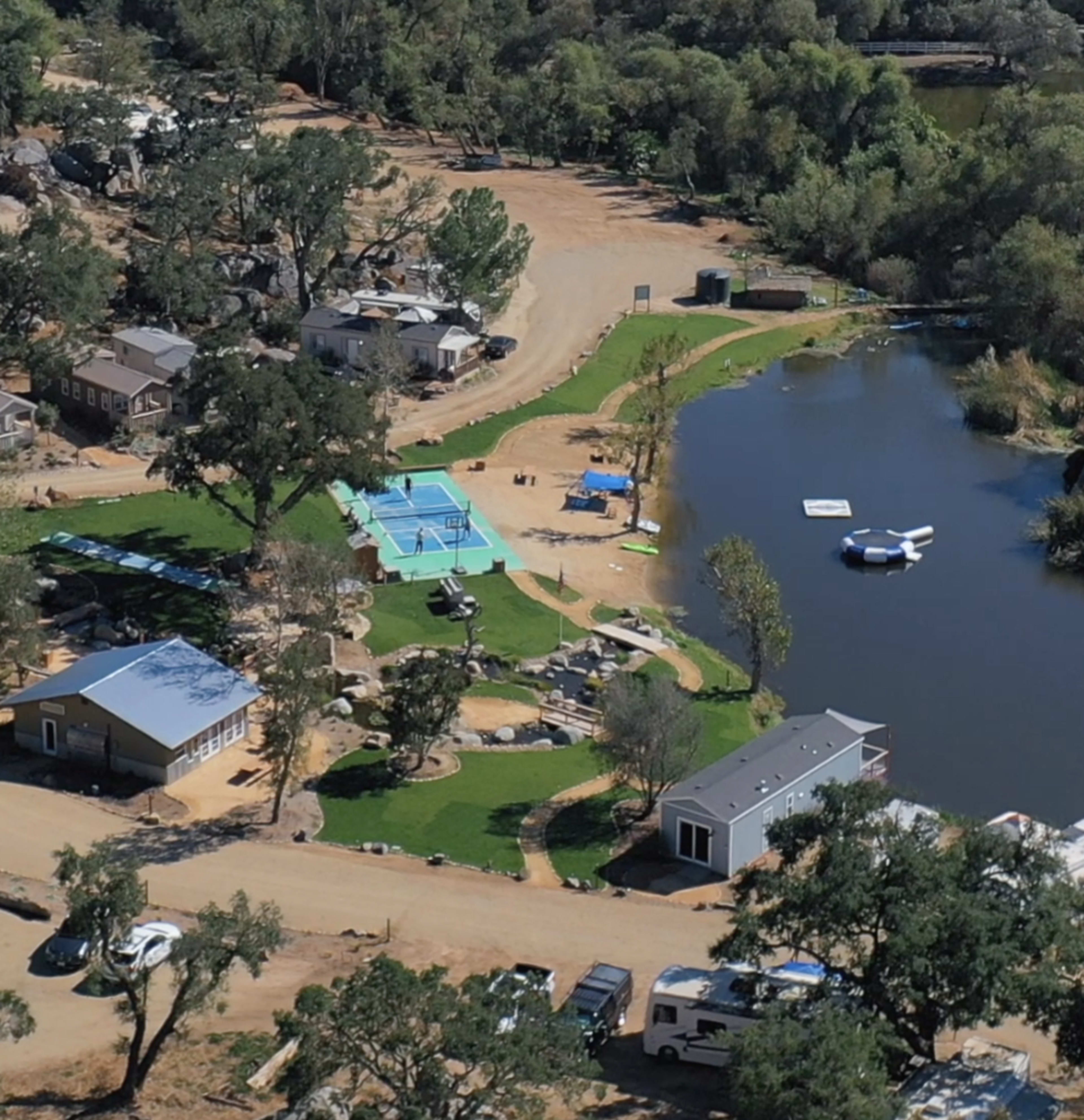 The image shows a recreational area featuring several houses, a swimming pool, grassy spaces, and a pond with floating structures.