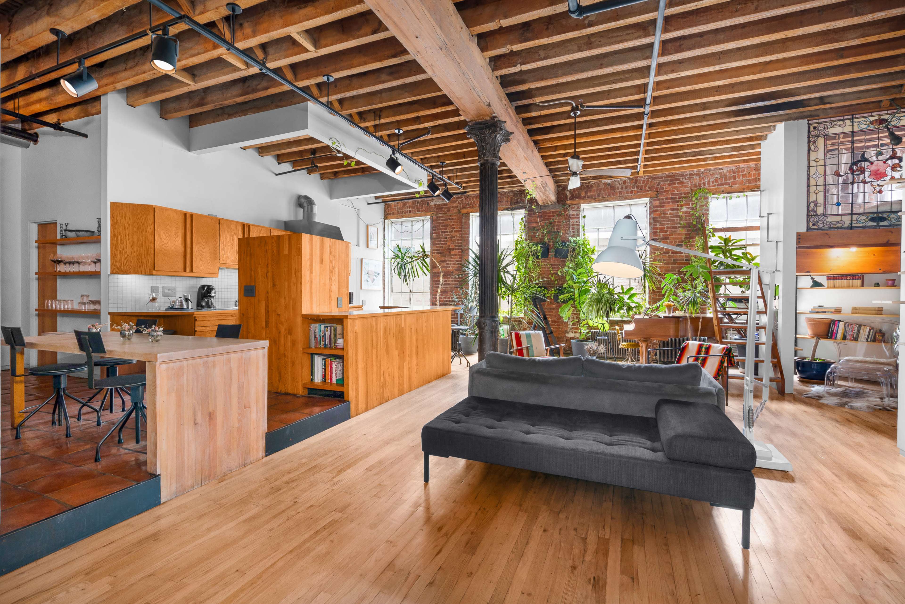 An open-concept living space featuring a kitchen with wooden cabinets, a seating area with a grey sofa, and large windows surrounded by plants.