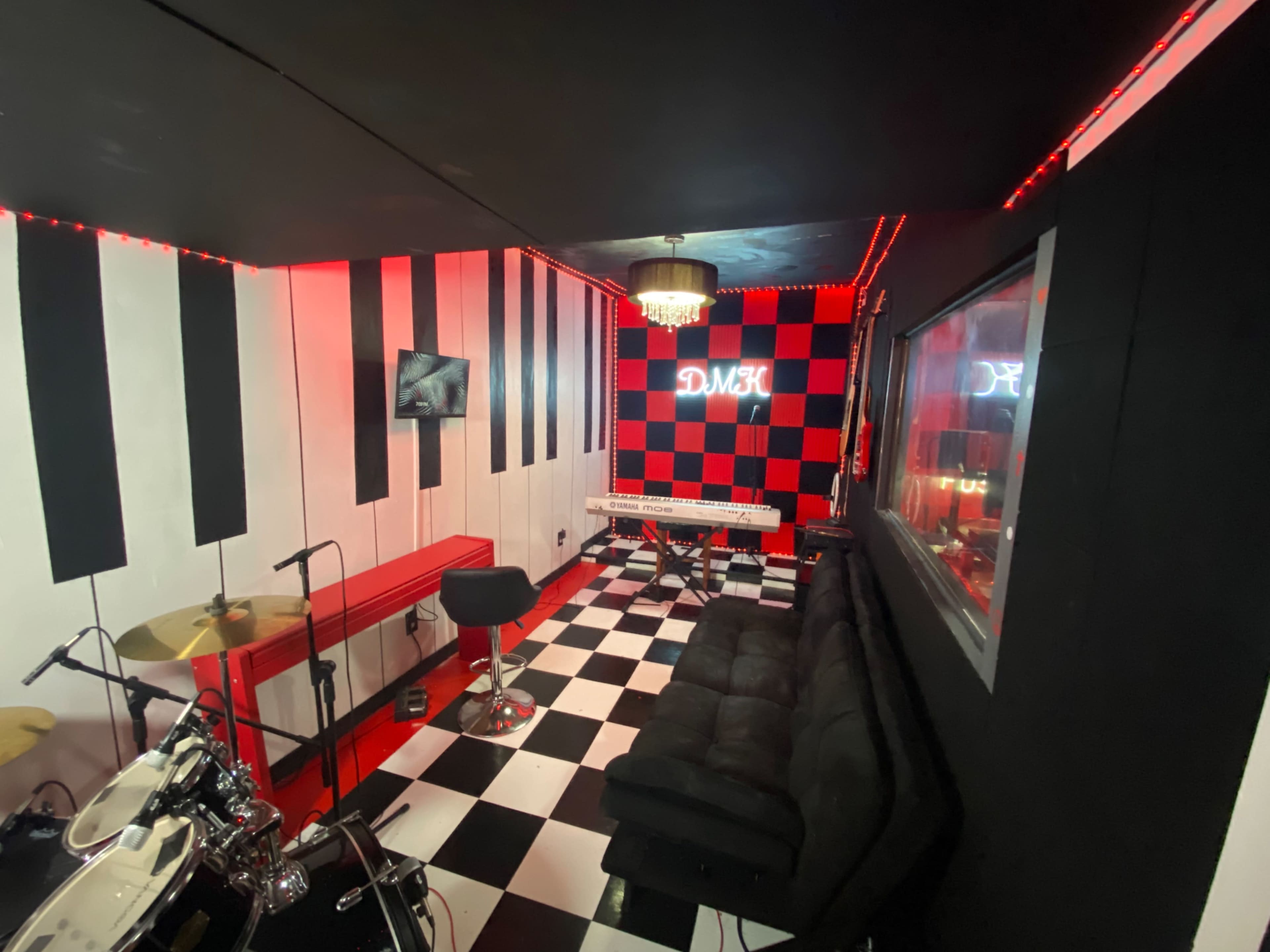 A music studio with a black and white checkered floor, red and black walls, a keyboard, a drum set, and a couch arranged under decorative lighting.