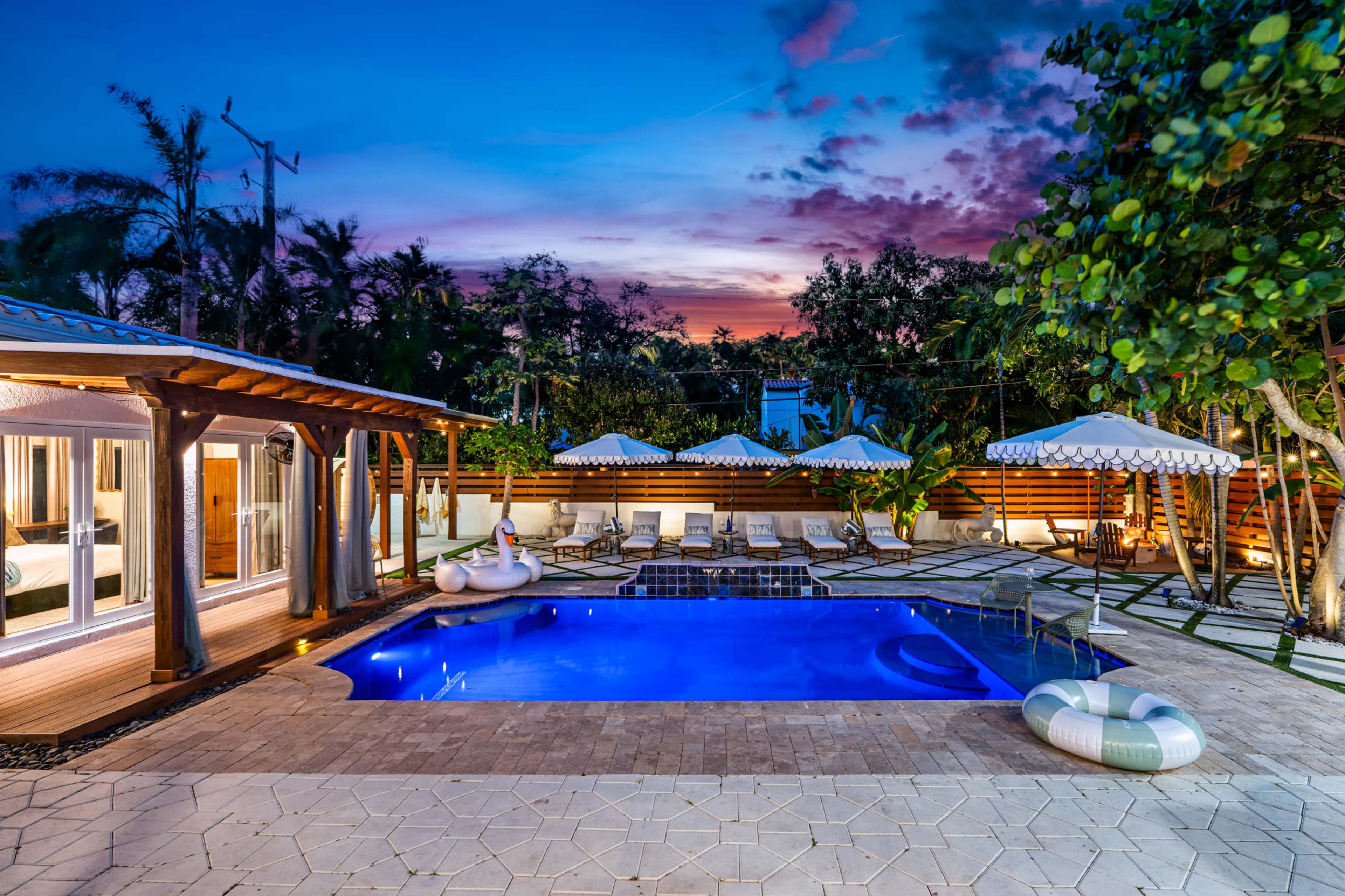 The image shows a well-lit outdoor pool area with lounge chairs and umbrellas, surrounded by lush greenery and a colorful sunset sky.