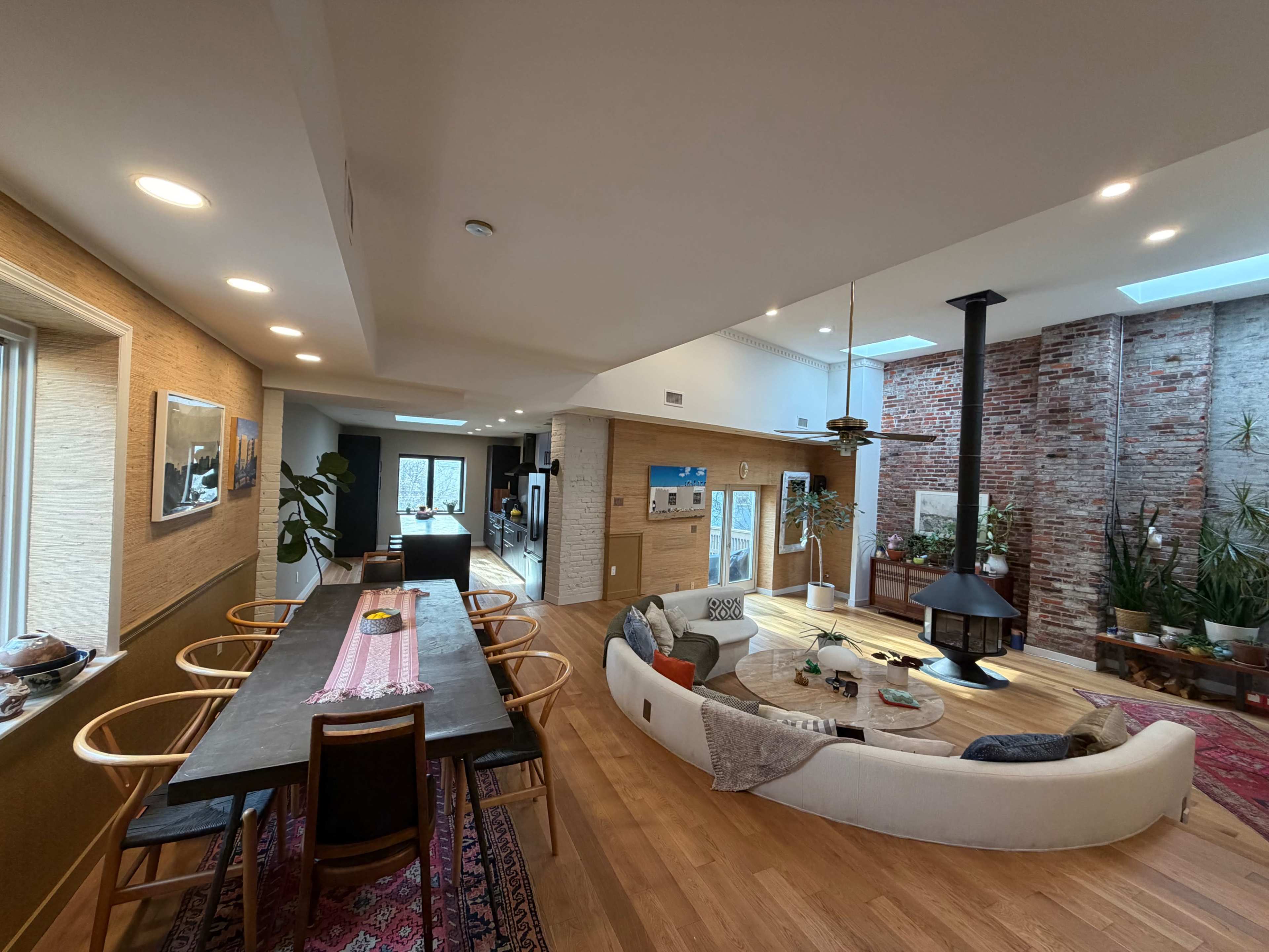 The image shows a spacious, modern living area featuring a dining table, a circular sofa, a fireplace, and large windows with natural light illuminating the open layout.