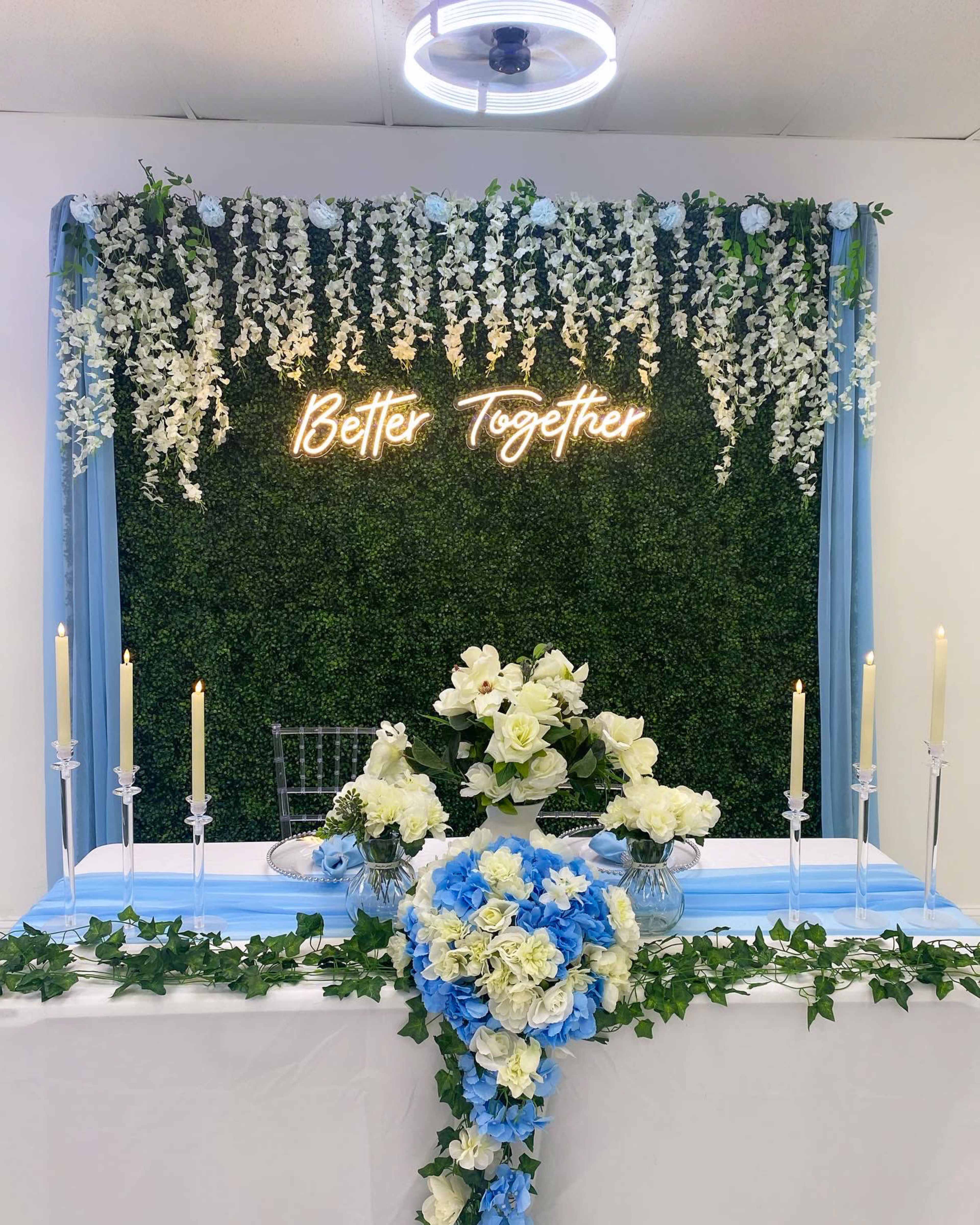A decorated wedding table is set against a floral backdrop with the phrase "Better Together" illuminated in neon lights.
