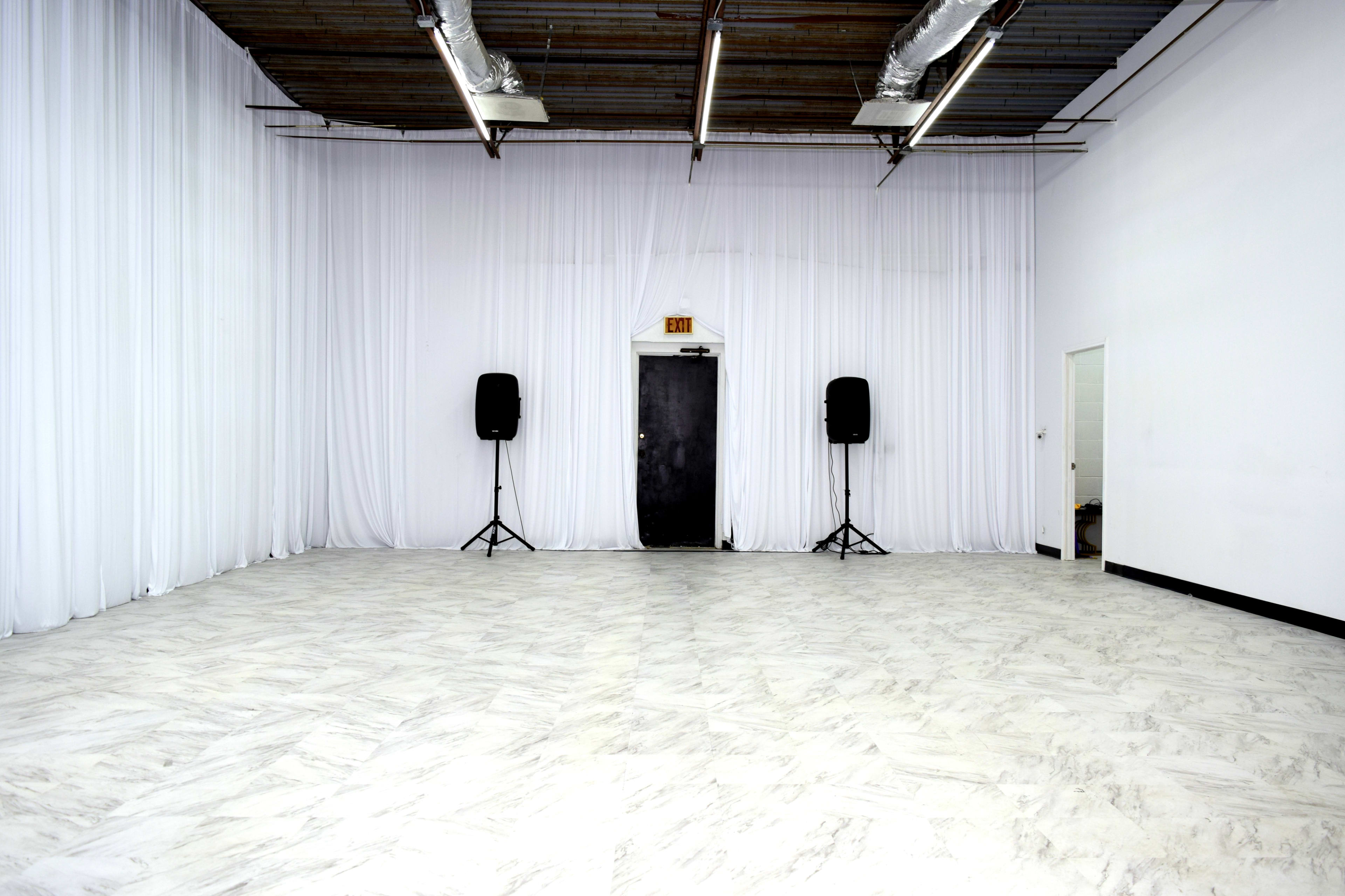 The image shows a large, empty room with white drapes, a black door, and two speaker stands positioned on either side.