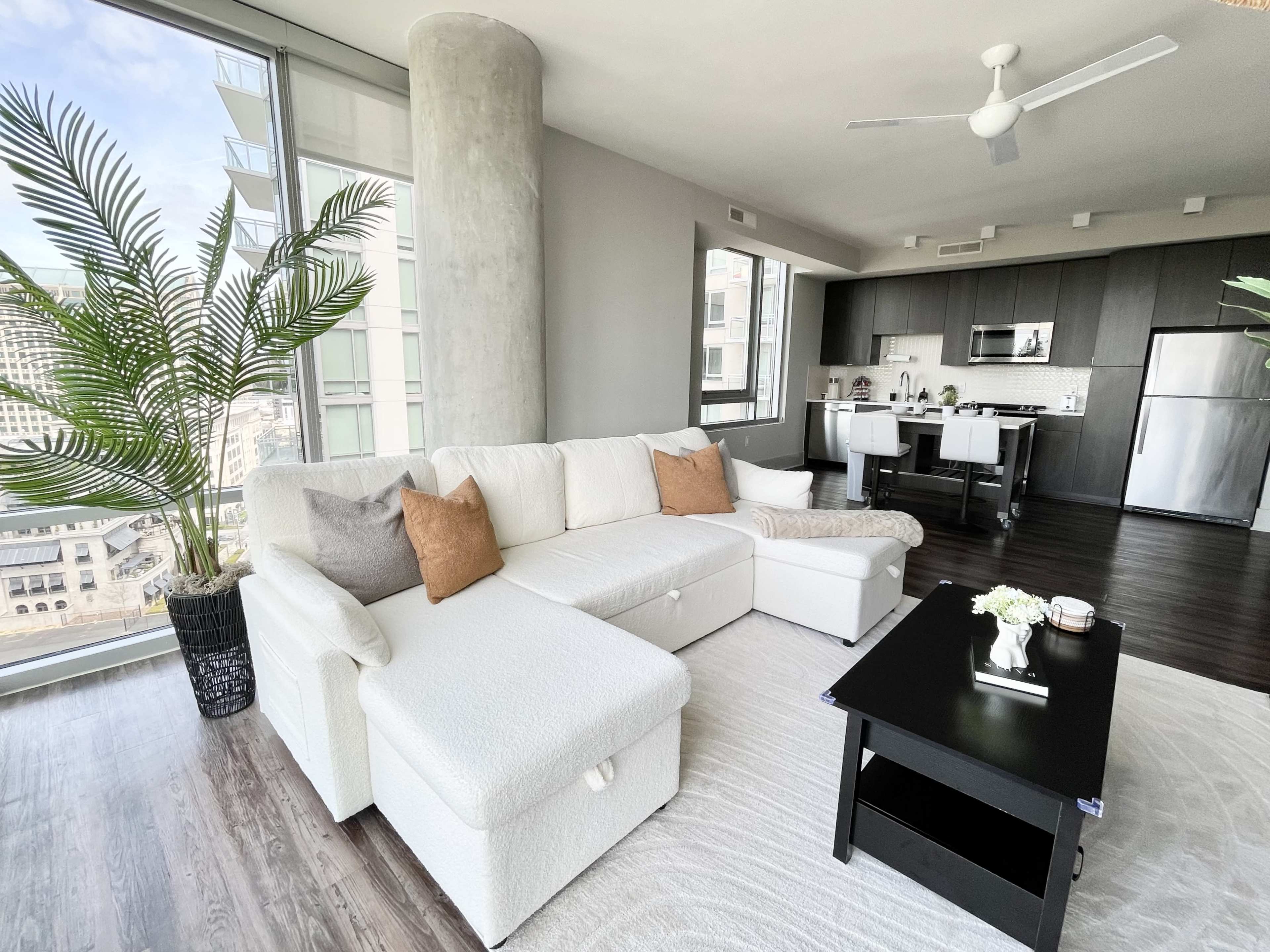 A modern living room features a white sectional sofa, a black coffee table, and large windows with views of the city skyline.