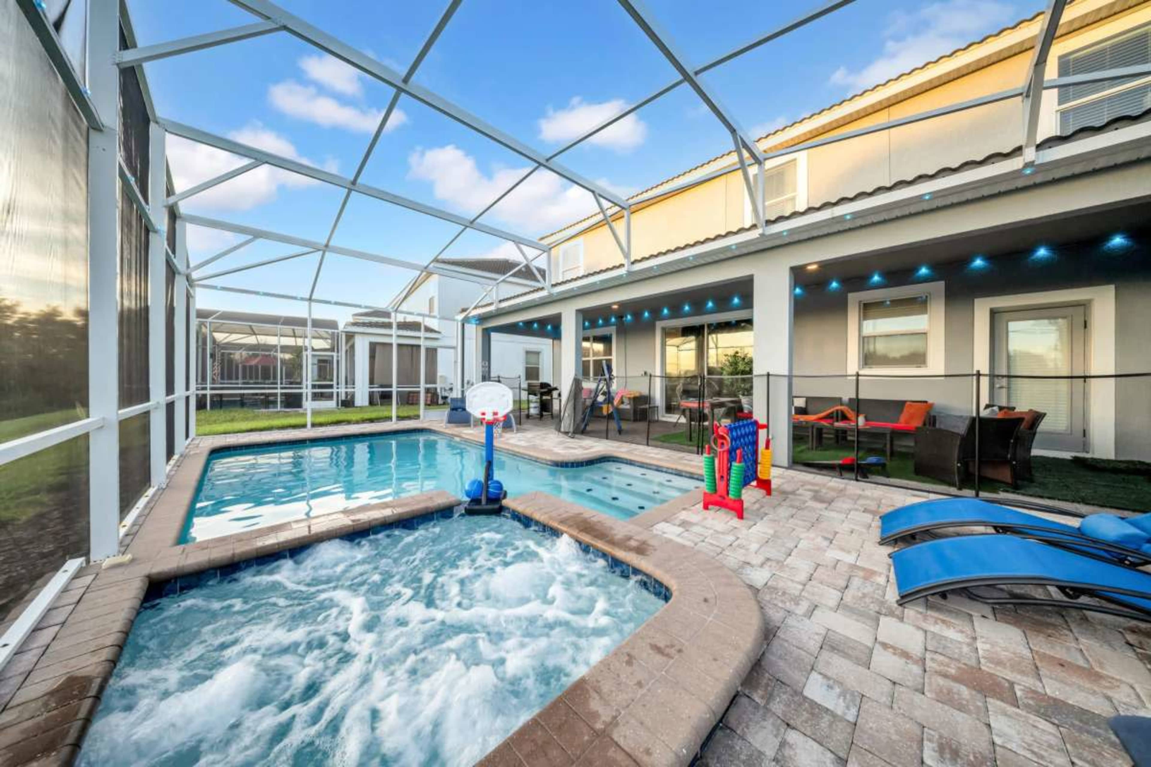 The image shows a fenced backyard with a swimming pool, a hot tub, a basketball hoop, lounge chairs, and outdoor seating under a screened enclosure.