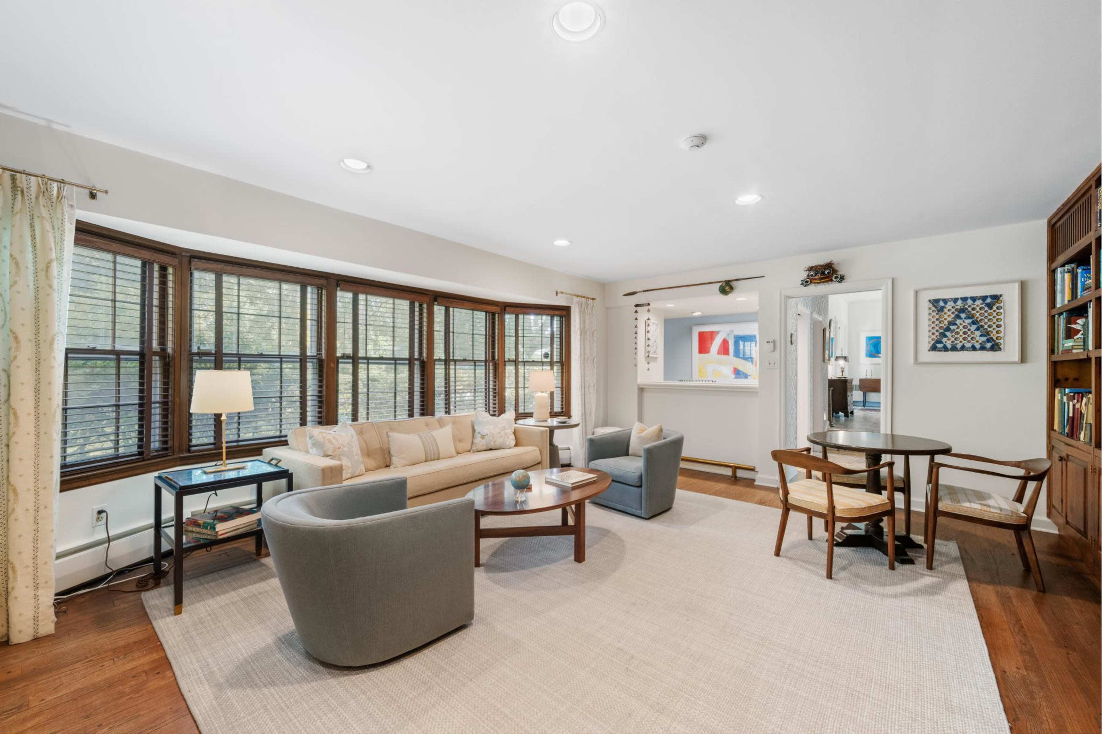 The image shows a living room with large windows, a beige couch, two gray armchairs, a round dining table, and a built-in bookshelf.