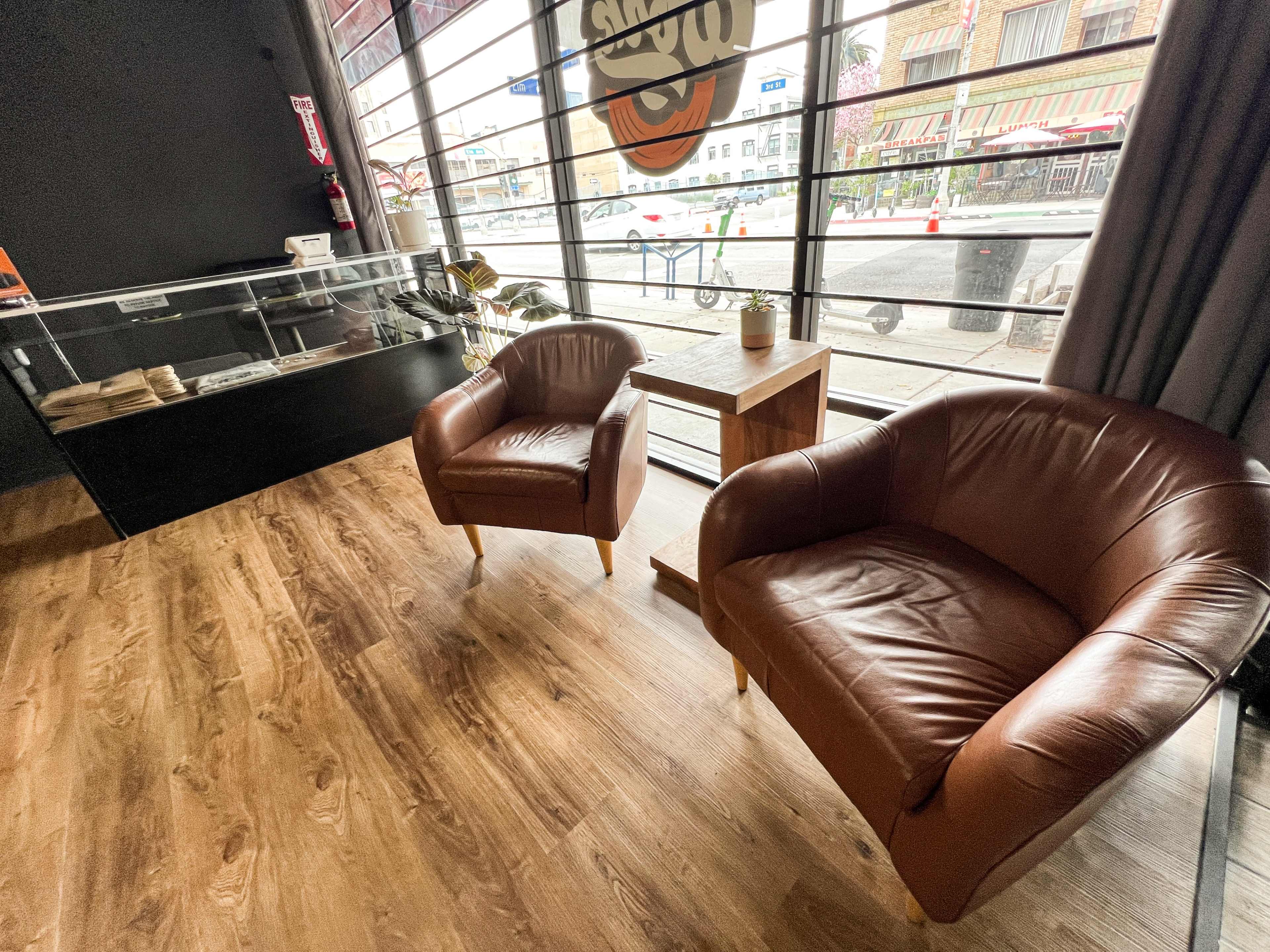 A coffee shop interior with two brown leather chairs facing a wooden table, large windows, and a glass display case in the background.