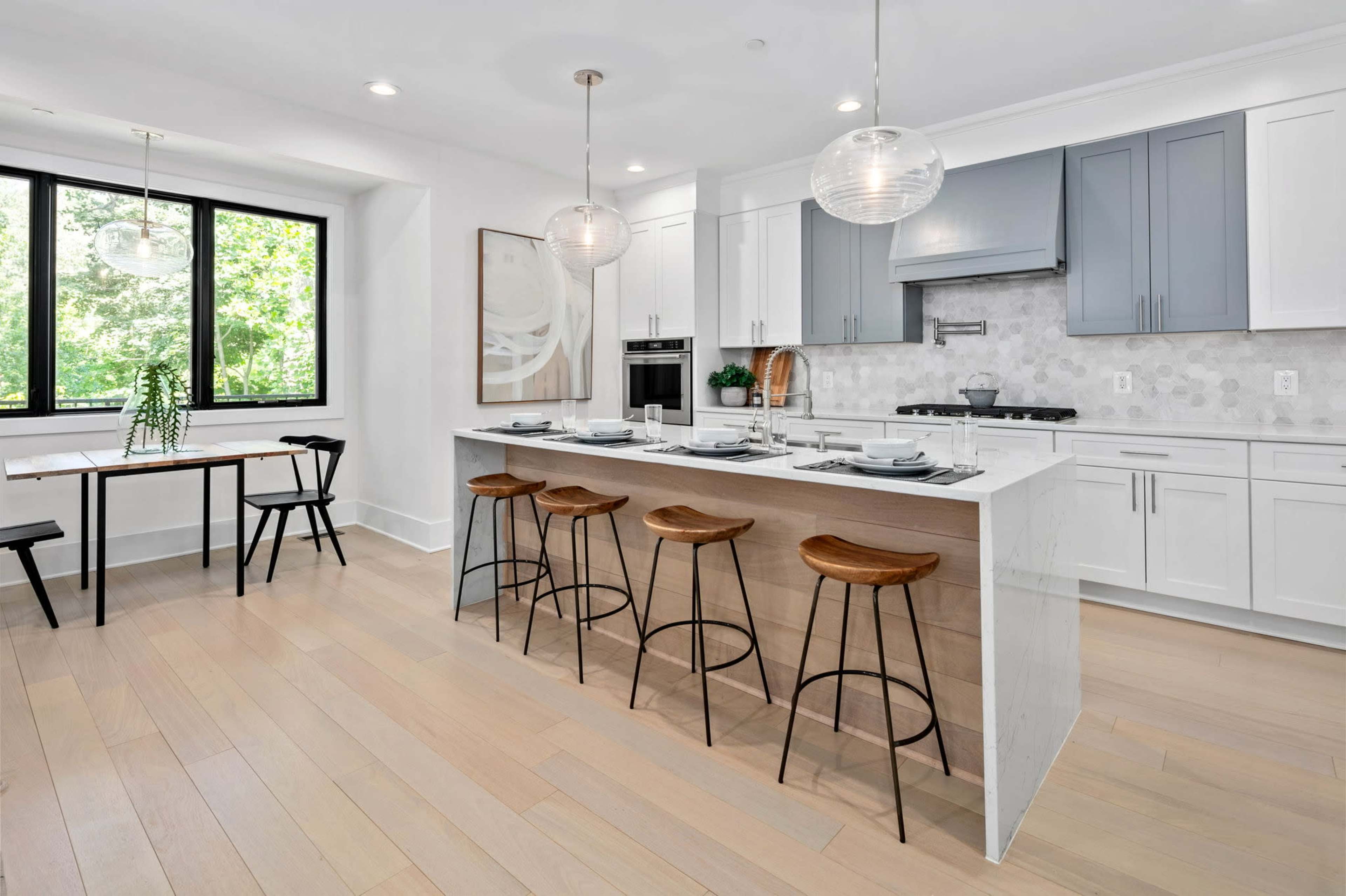 A modern kitchen with a large island, four bar stools, and a dining table near a window overlooking greenery.