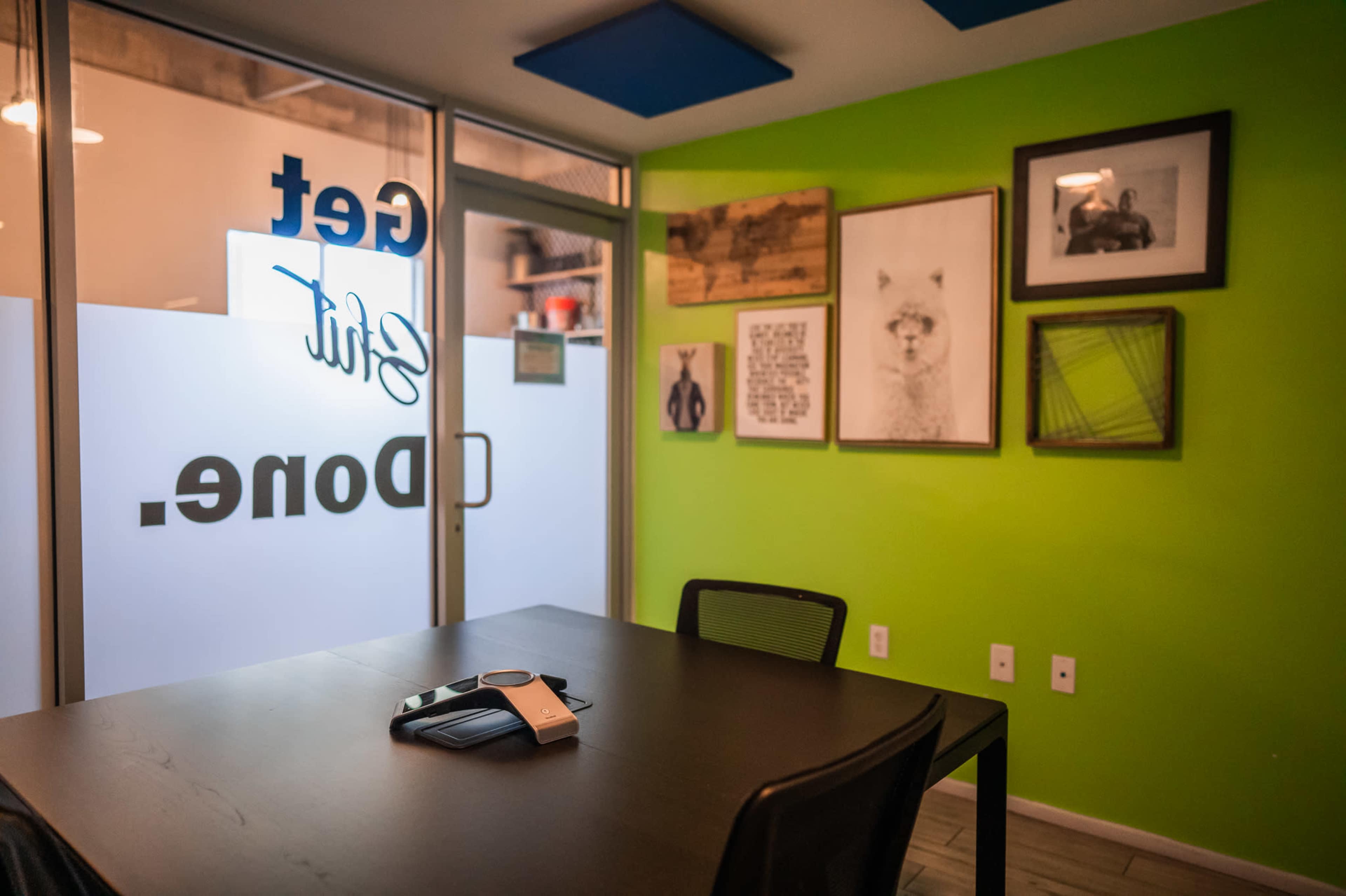 A meeting room features a black table with a phone and several framed artworks on bright green walls.