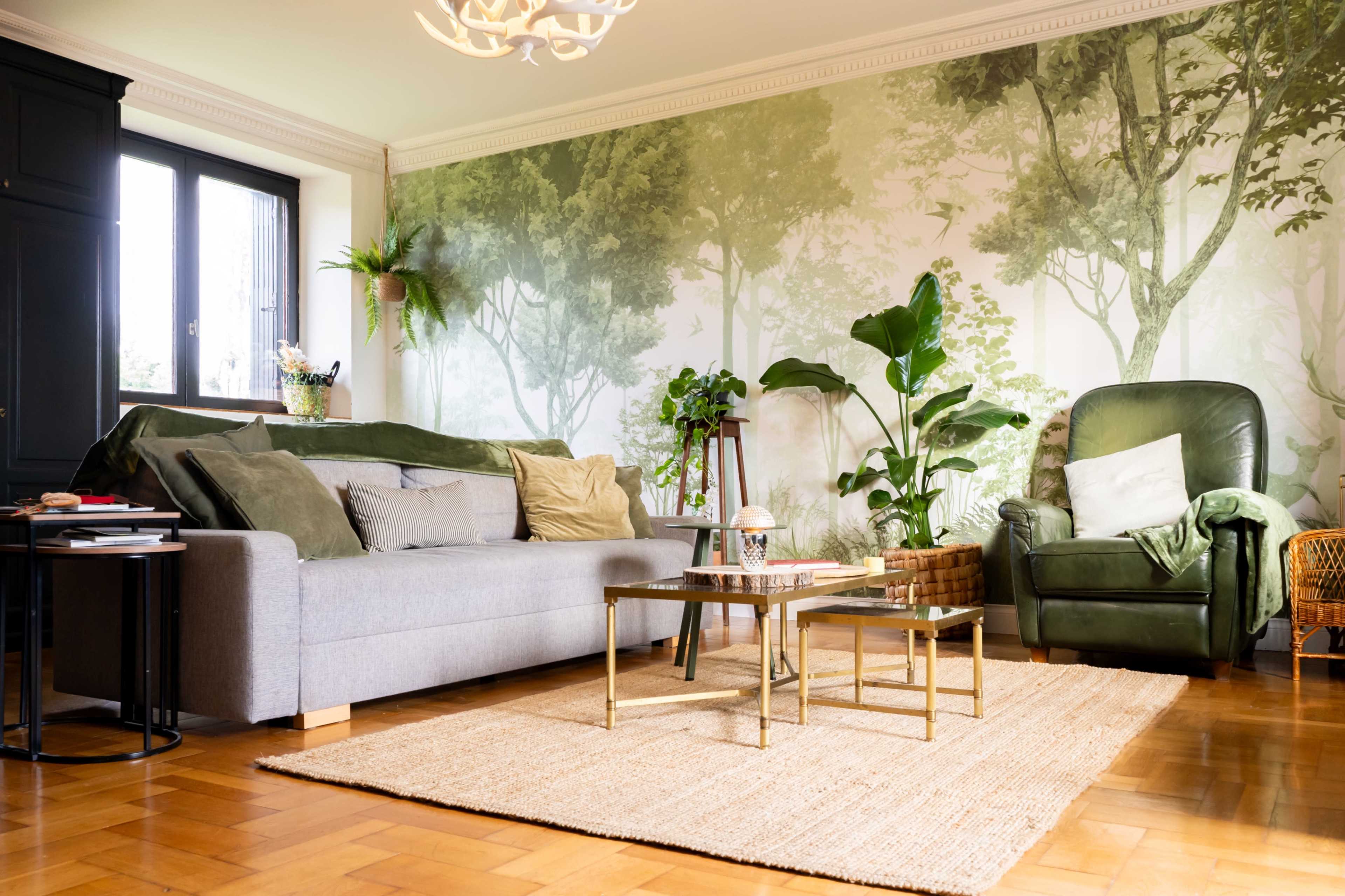 A cozy living room features a gray sofa and a green armchair, surrounded by plants and a large wall mural of trees.