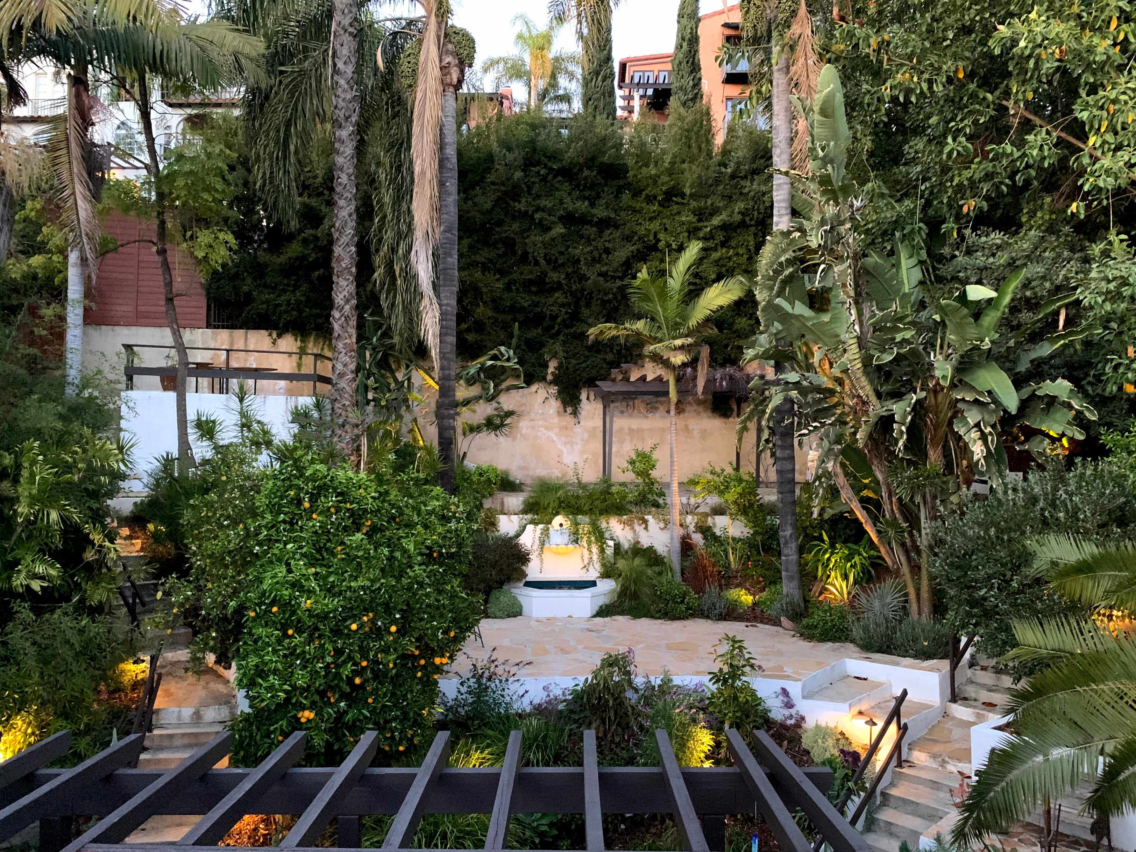 The image depicts a landscaped garden with various plants and palm trees, featuring a small fountain at the center surrounded by stone pathways.