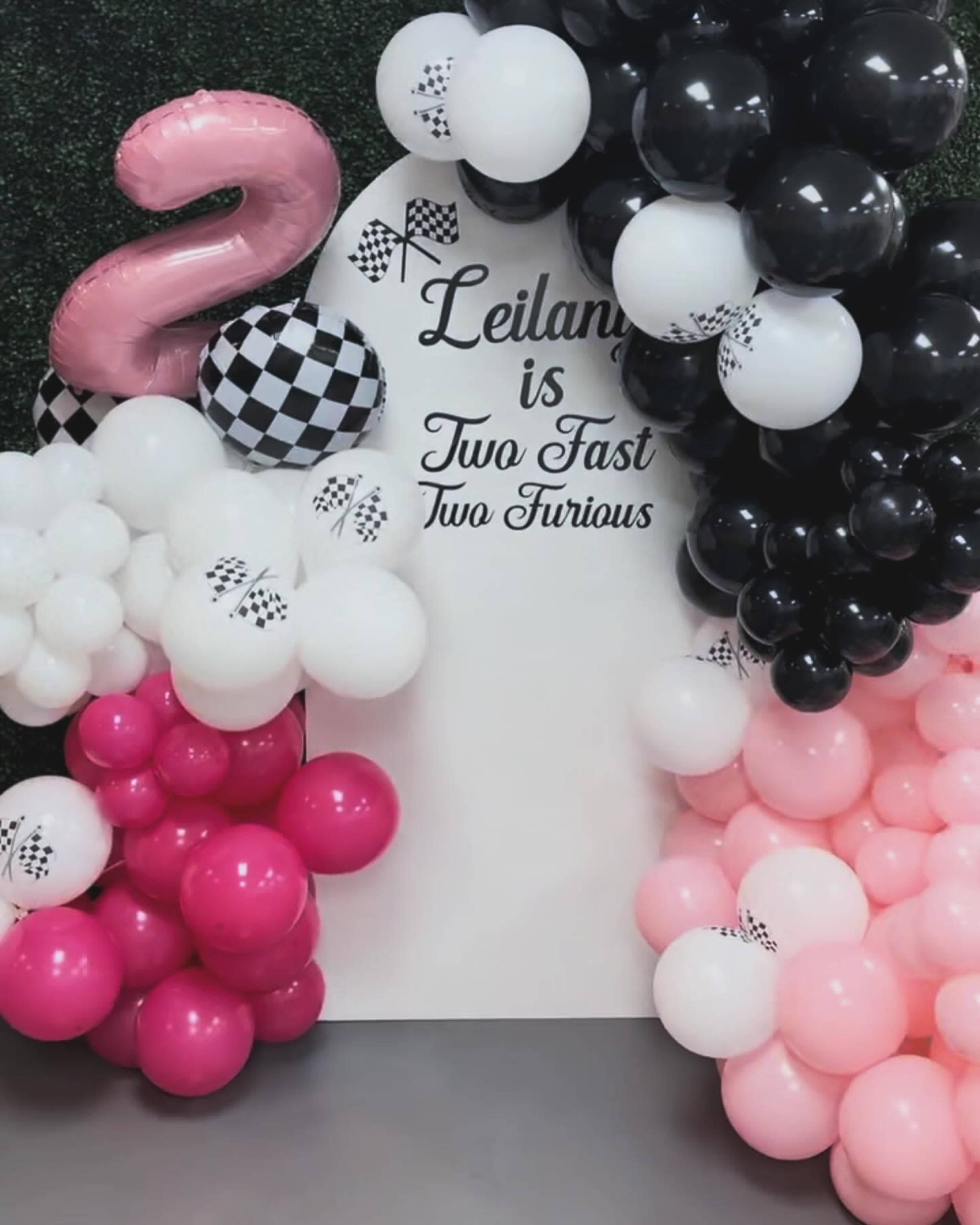 The scene features a decorated wall with a large "2" balloon and an assortment of pink, black, and white balloons arranged around the text "Leilani is Two Fast Two Furious."