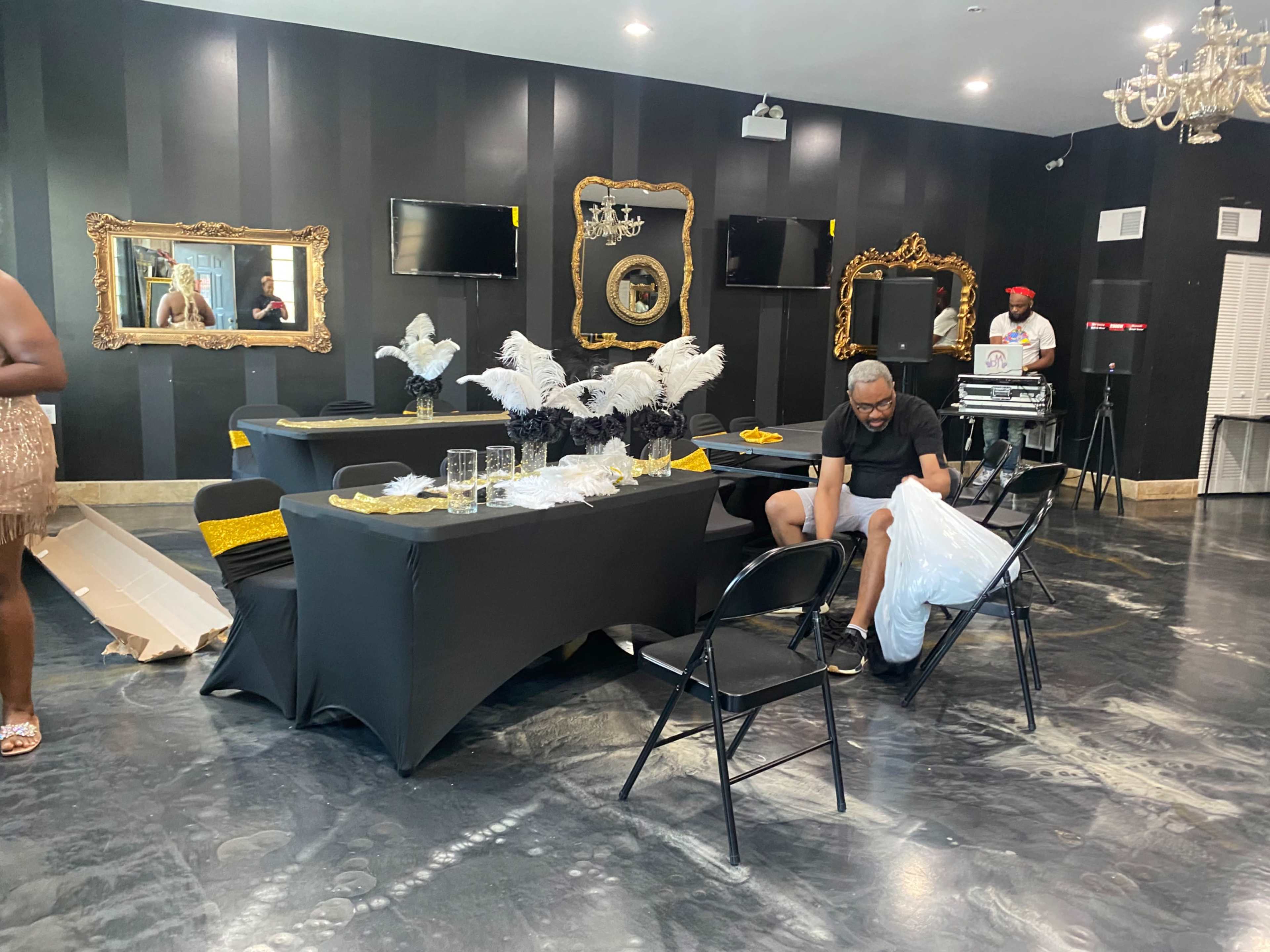 A decorated event space features black tables with gold accents, while a man in glasses adjusts a white chair in the foreground.