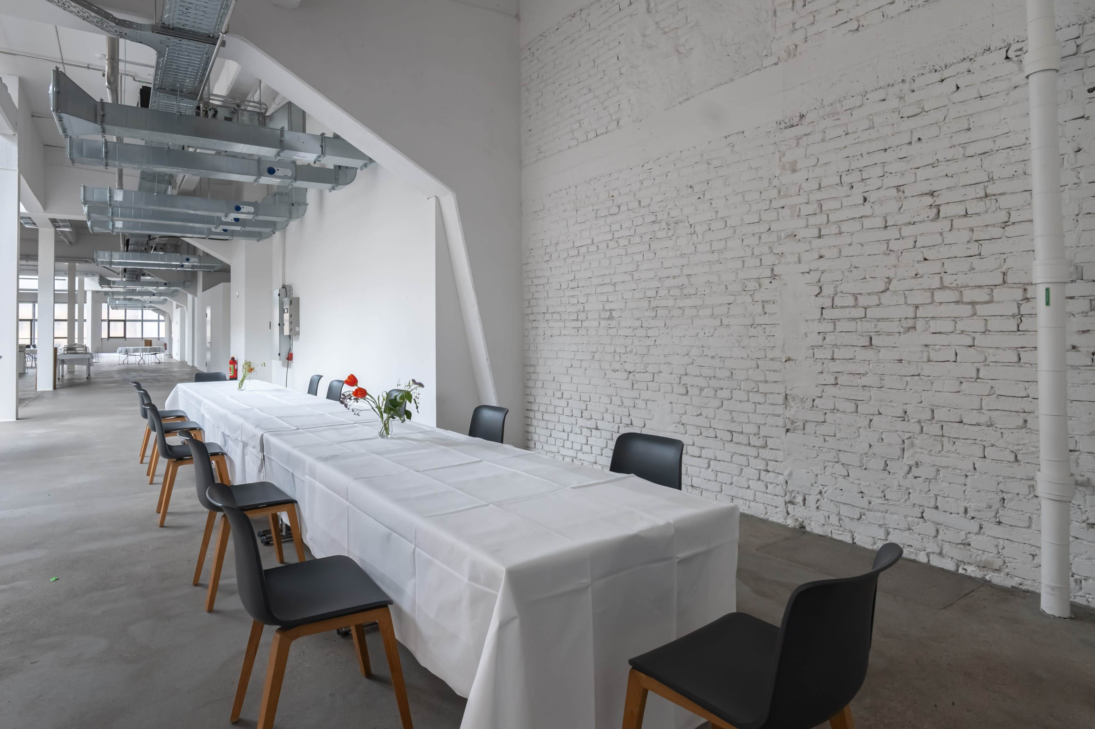 A long, white-covered table is set in a spacious room with a white brick wall and black chairs arranged around it.
