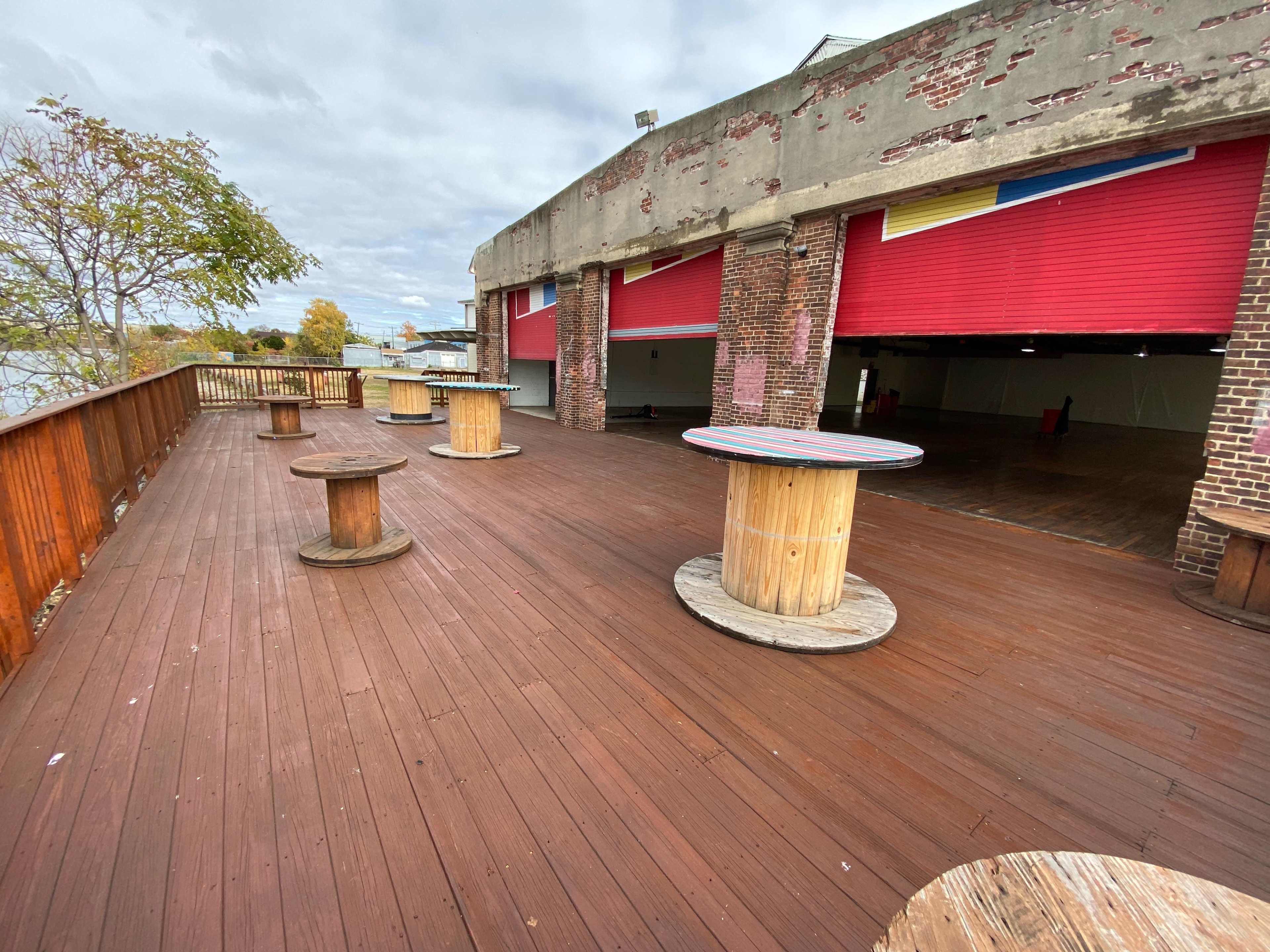 A wooden deck area with several round tables made from large cable spools, set against a backdrop of brick walls and red garage doors.
