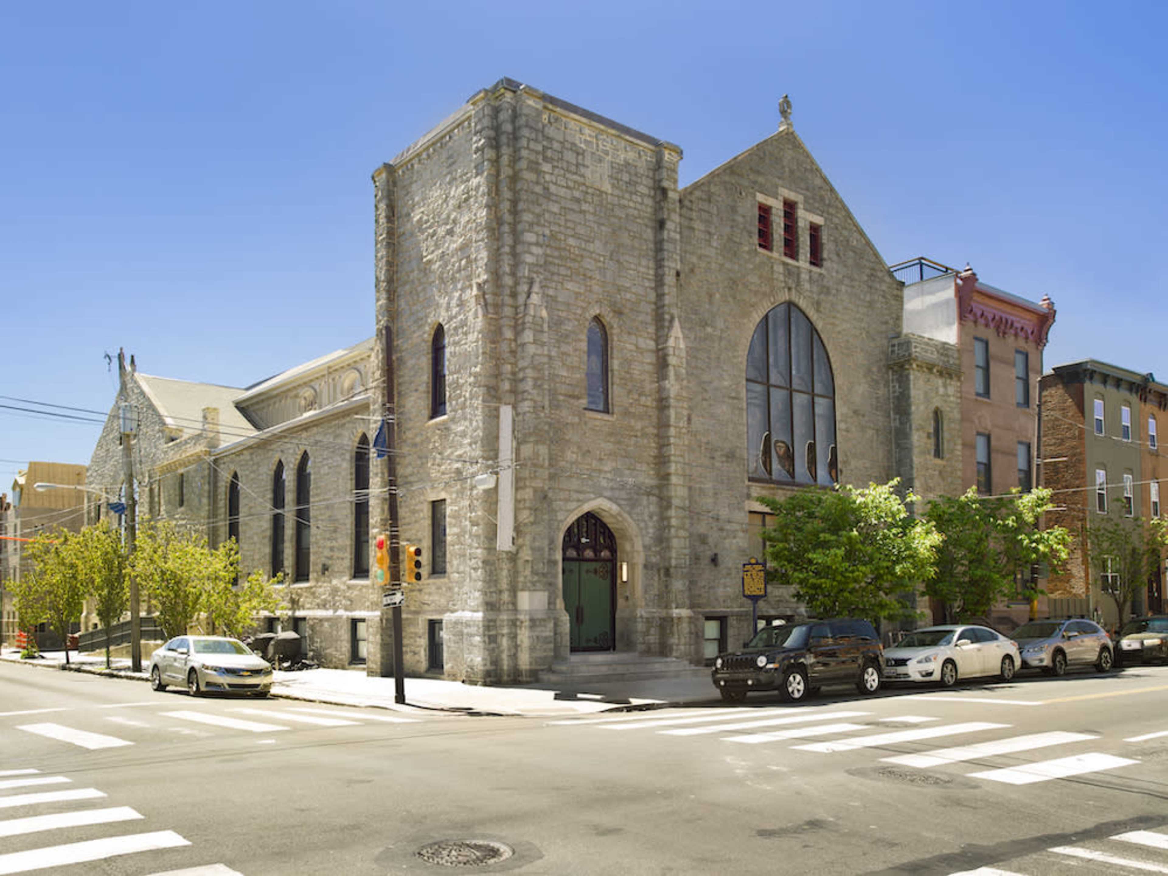 The image shows a stone church building at the intersection of two streets, surrounded by parked cars and trees under a clear blue sky.