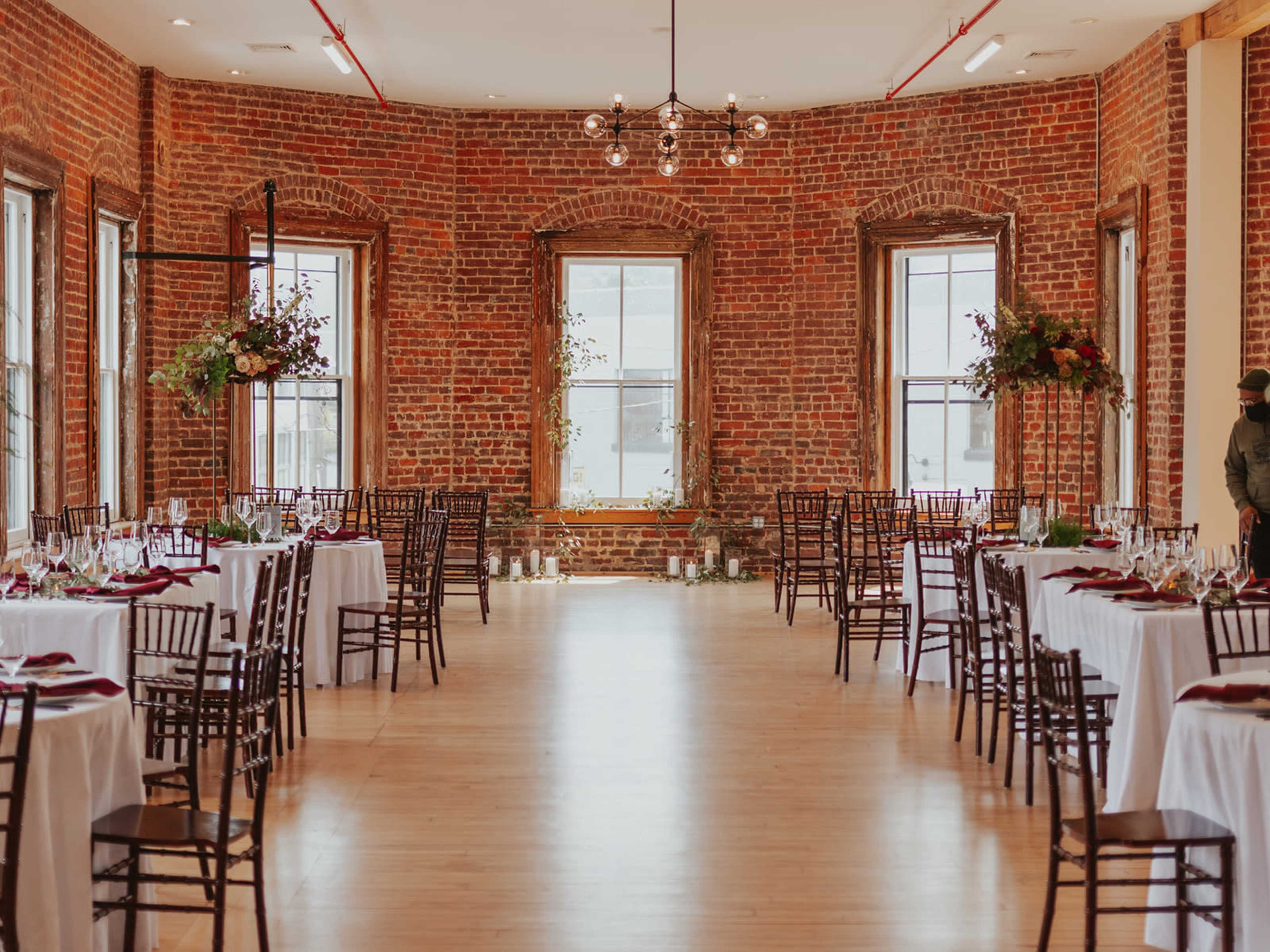 A spacious banquet hall features white tablecloths and dark wooden chairs arranged around a polished wooden floor, with exposed brick walls and large windows providing natural light.
