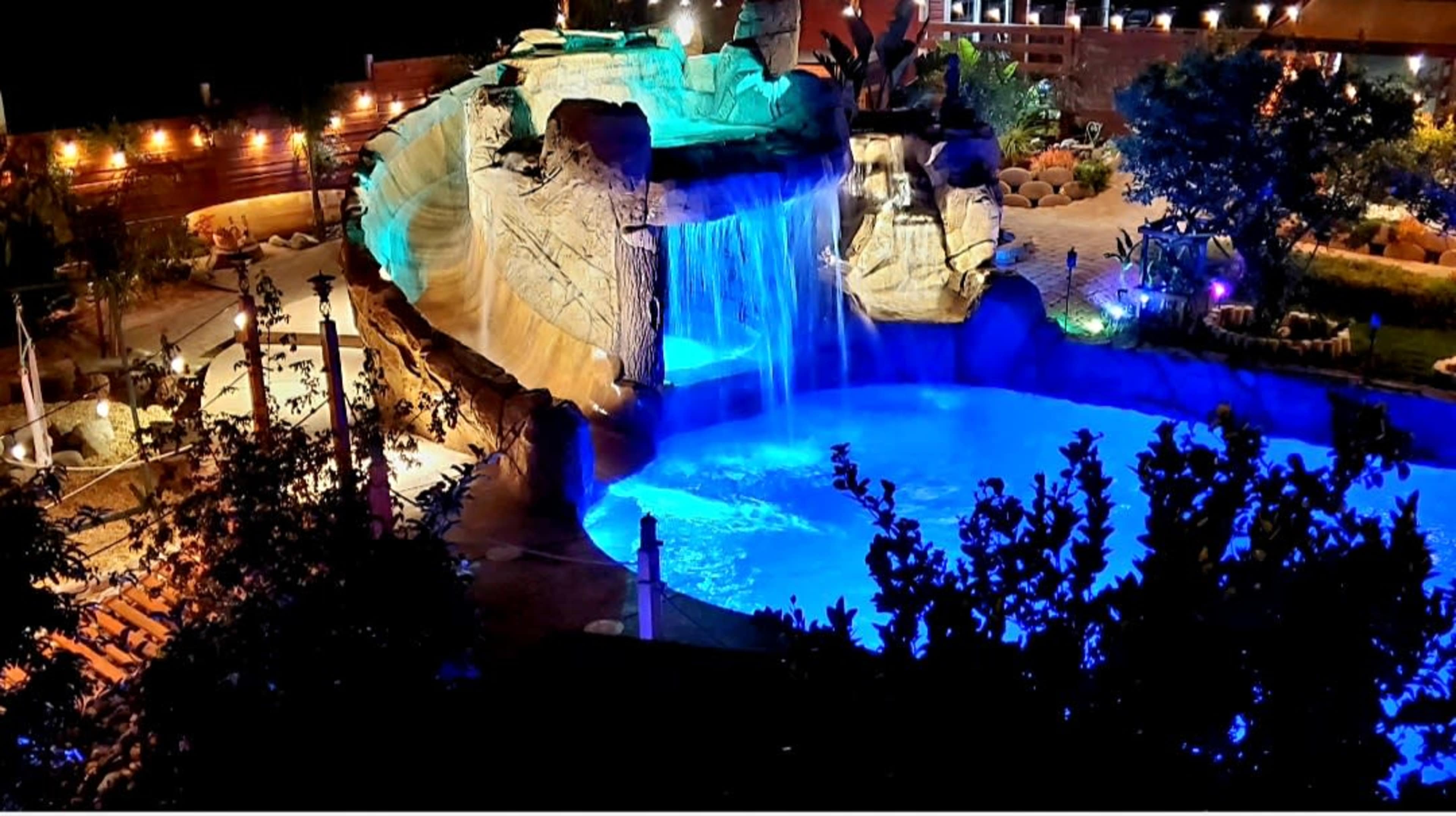 The image shows a brightly lit outdoor pool area with a waterfall cascading over a rock formation, surrounded by landscaping and illuminated by various lights.