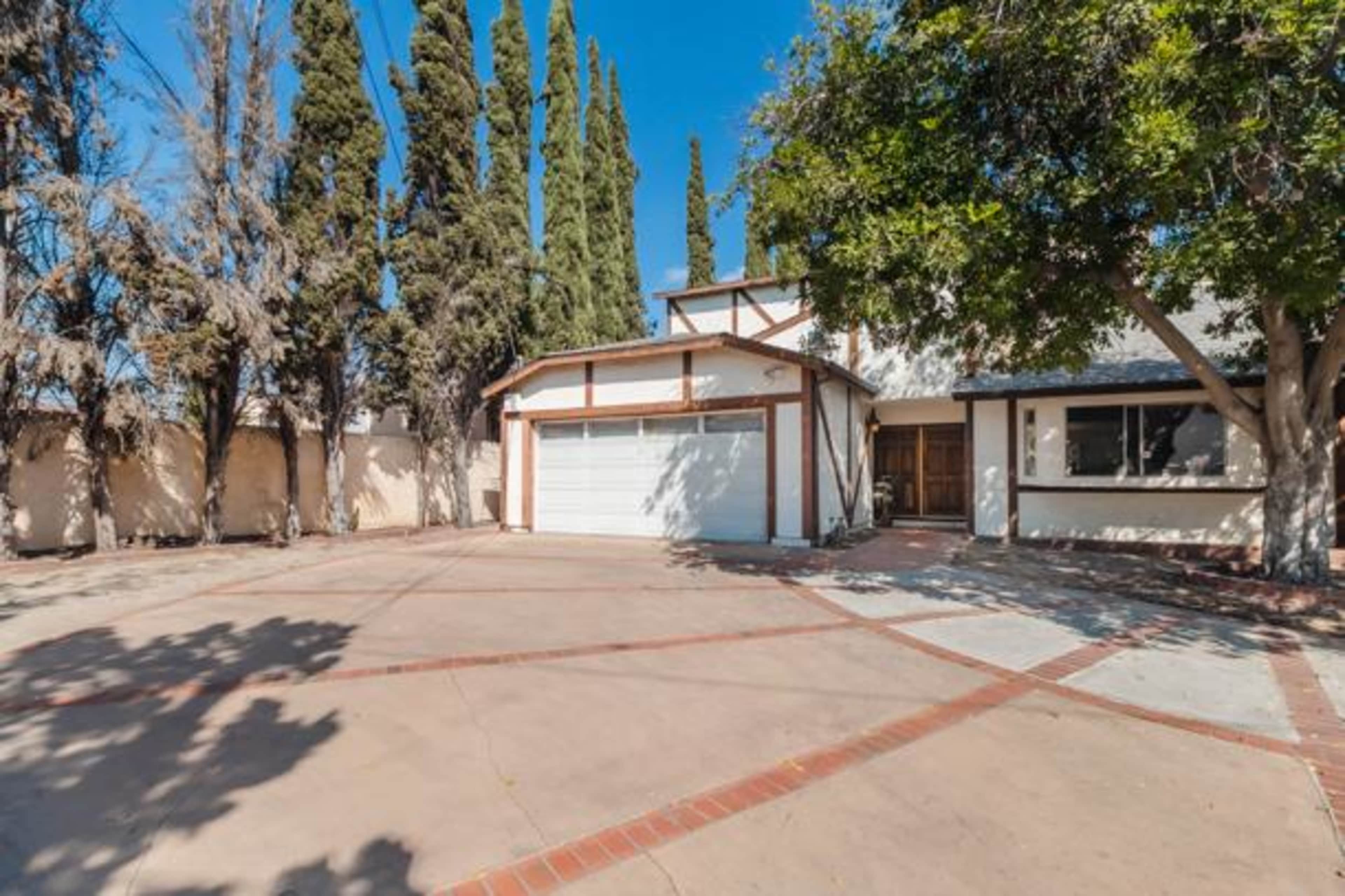 The image shows a two-story house with a large driveway surrounded by tall trees.
